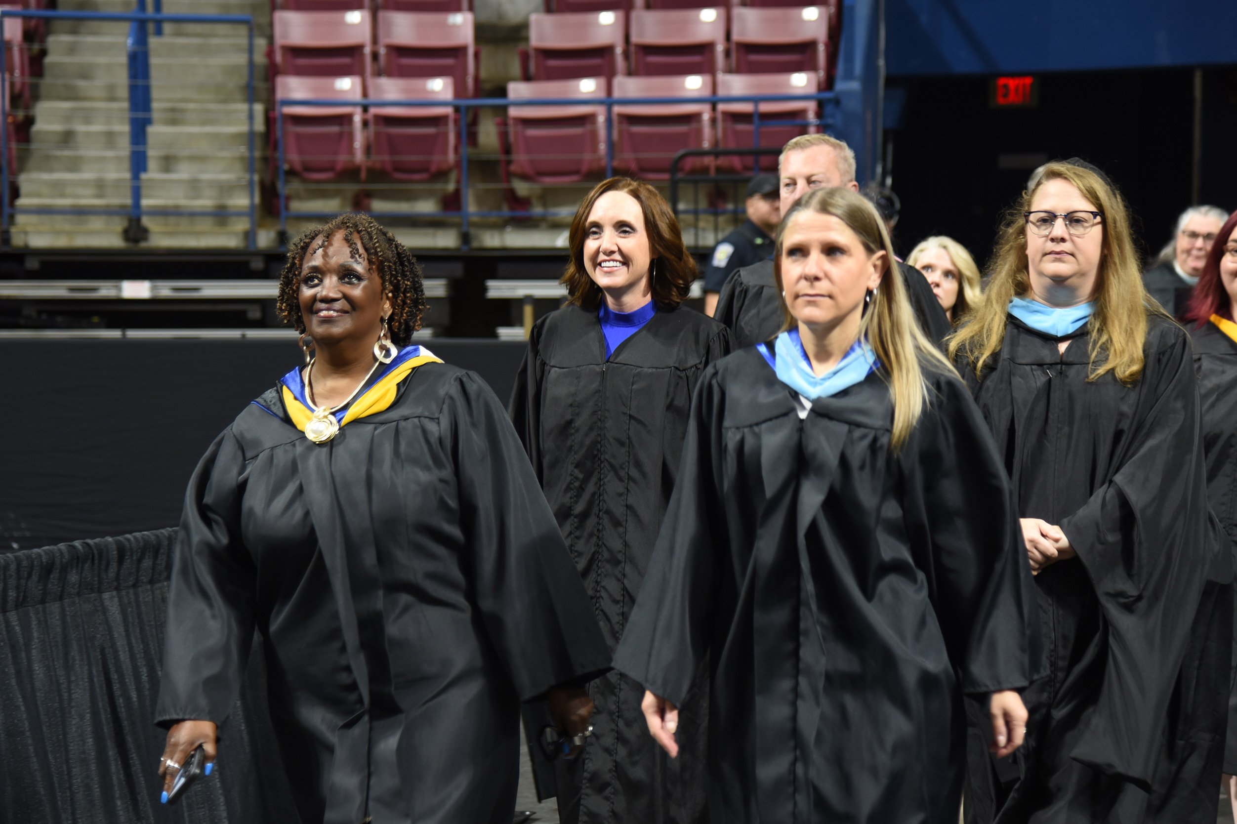 Five women in black gowns and medals walk together in a stadium. The rows of seats are empty.