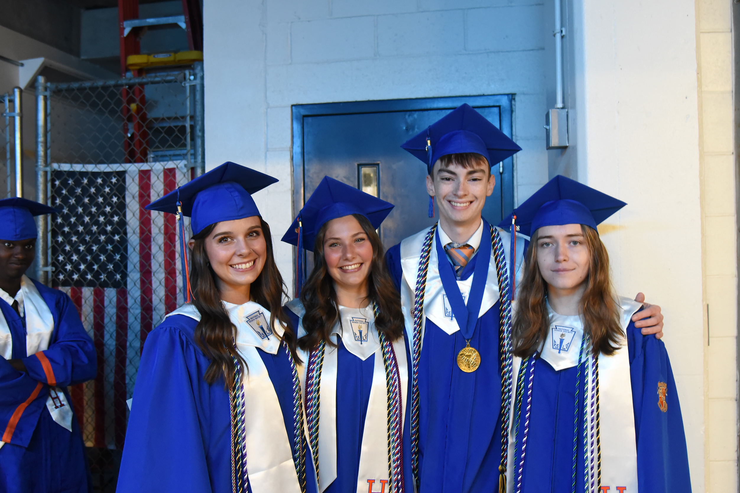 Five people in graduation caps and gowns pose for a picture in front of a wall and an American flag.