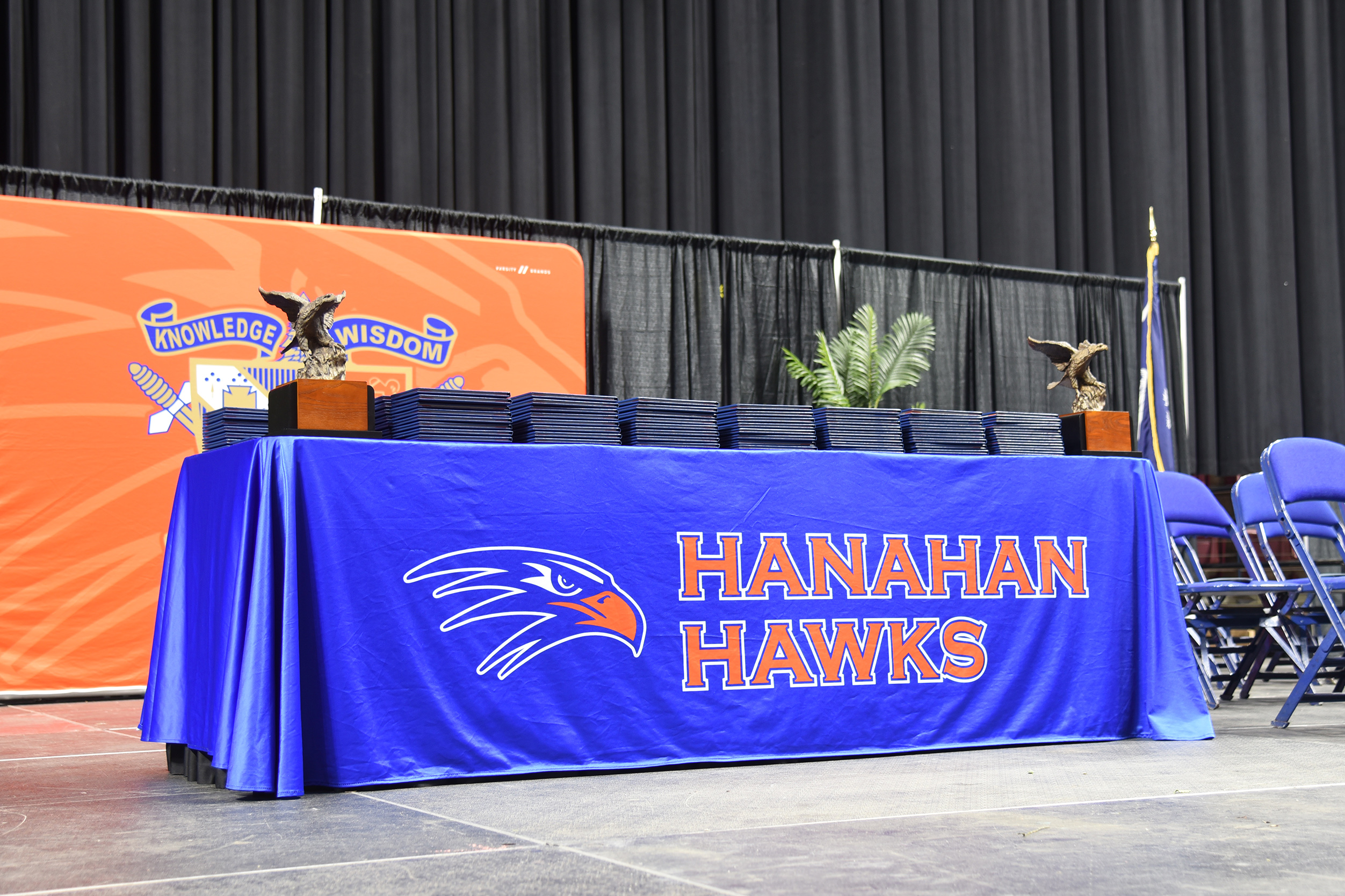 Blue table with "Hanahan Hawks" logo. Two trophies on the table, a potted plant behind it, and black curtains in the background.
