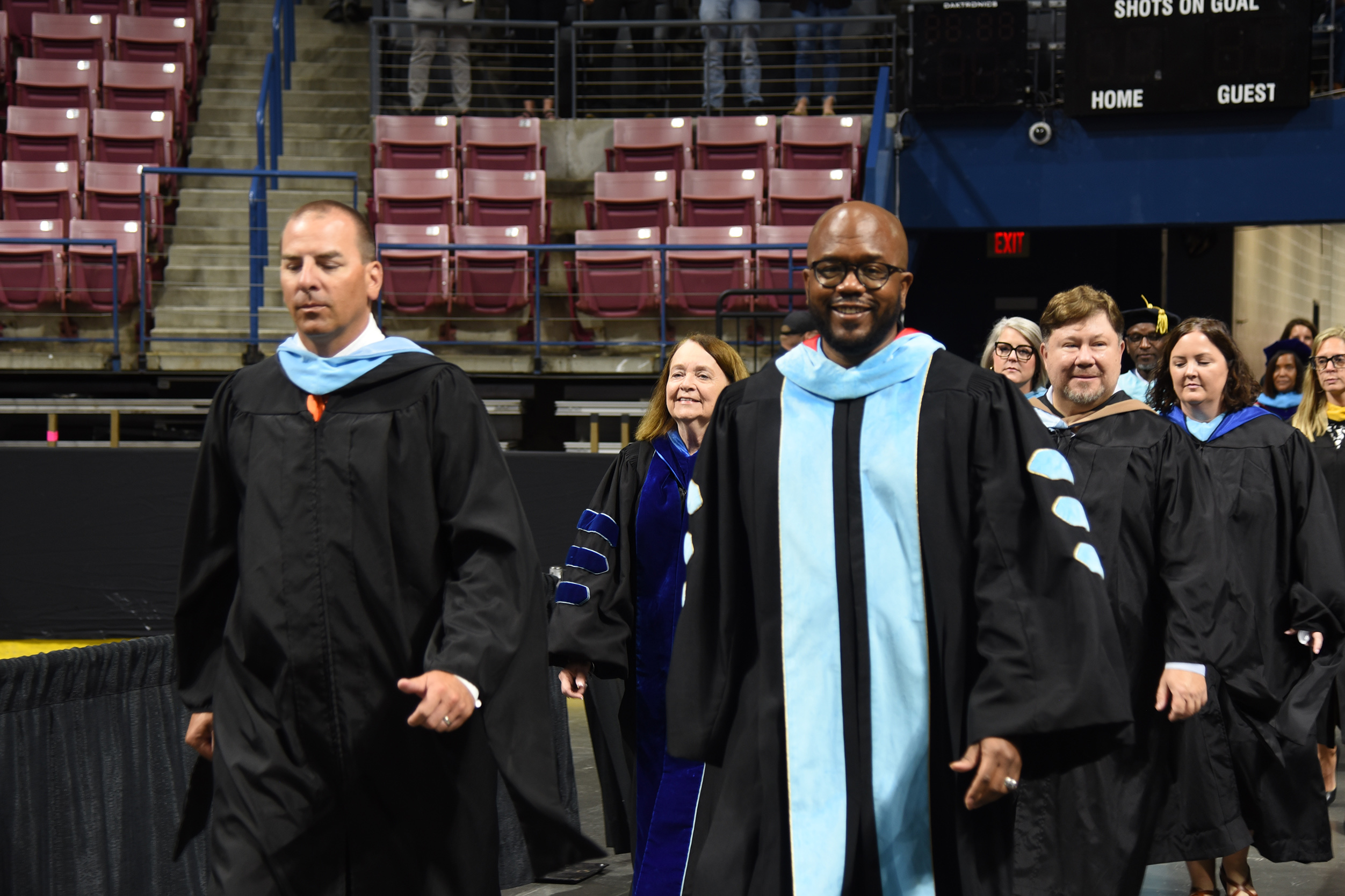 Graduates in black gowns and blue sashes walk in an indoor auditorium with empty seats in the background.