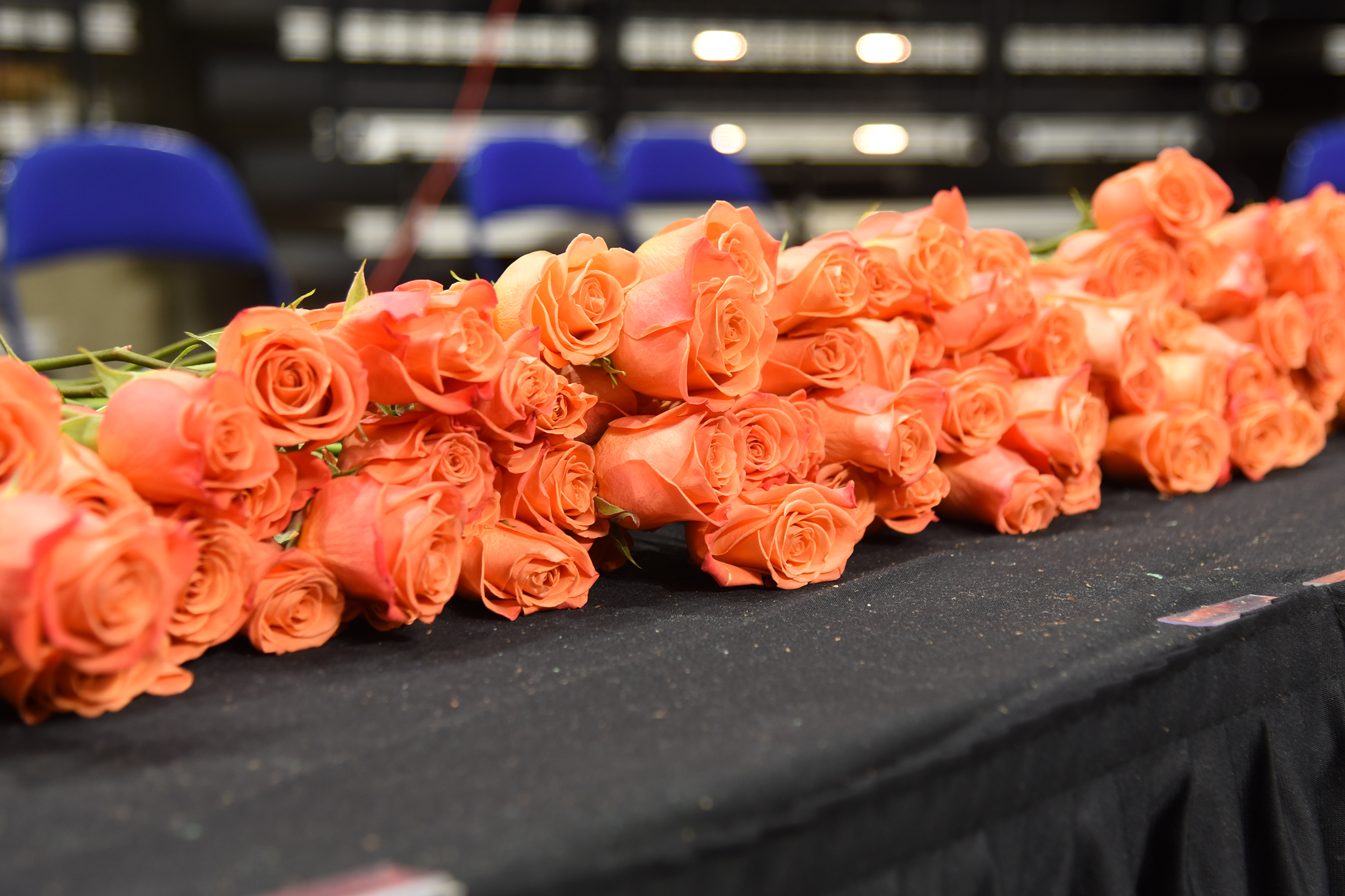A table with black cloth holds a row of orange roses. Three blue chairs are in the background.