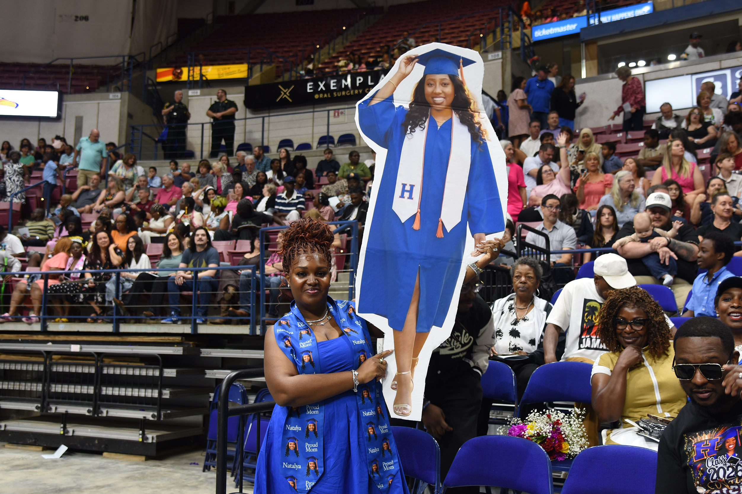 A woman holds a cutout of a graduate in a cap and gown at a sports event.