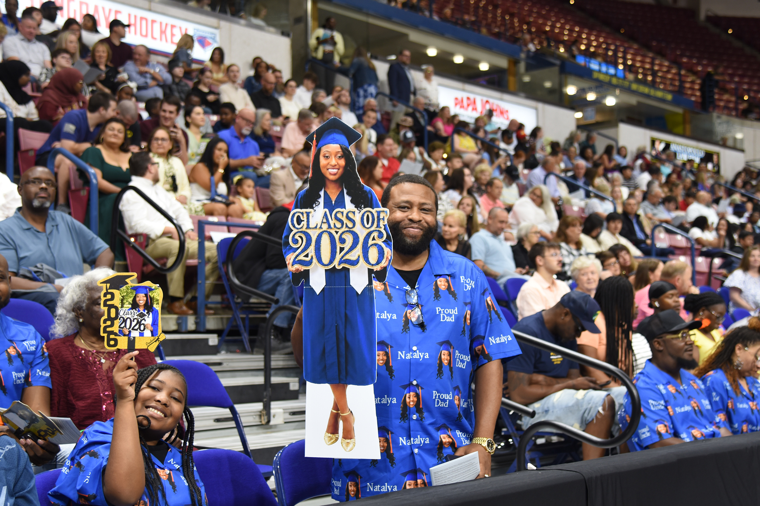 Audience seated in a stadium. Two people hold signs reading "Class of 2026" and "2026" in front of them.