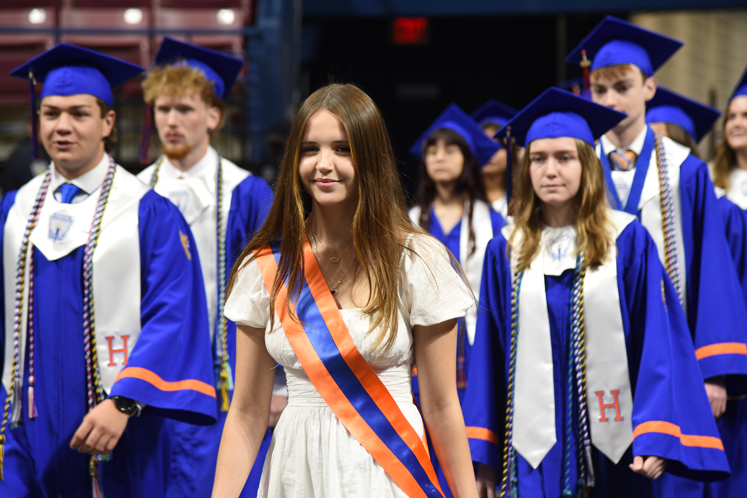 Graduates in blue and white robes and caps, a woman in a white dress with an orange sash.