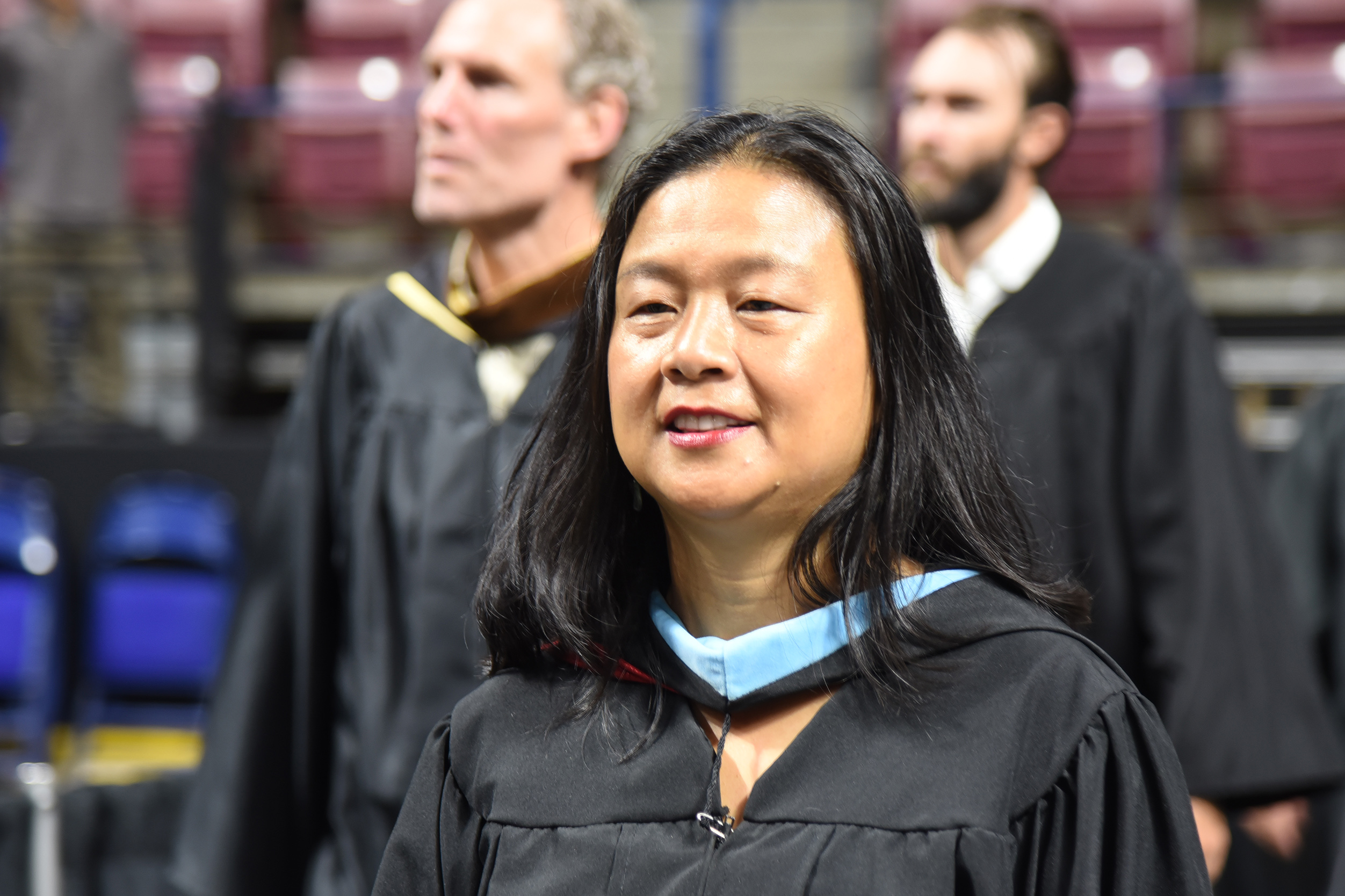 A woman in a black graduation gown with a blue stole smiles, standing with others in a graduation ceremony.