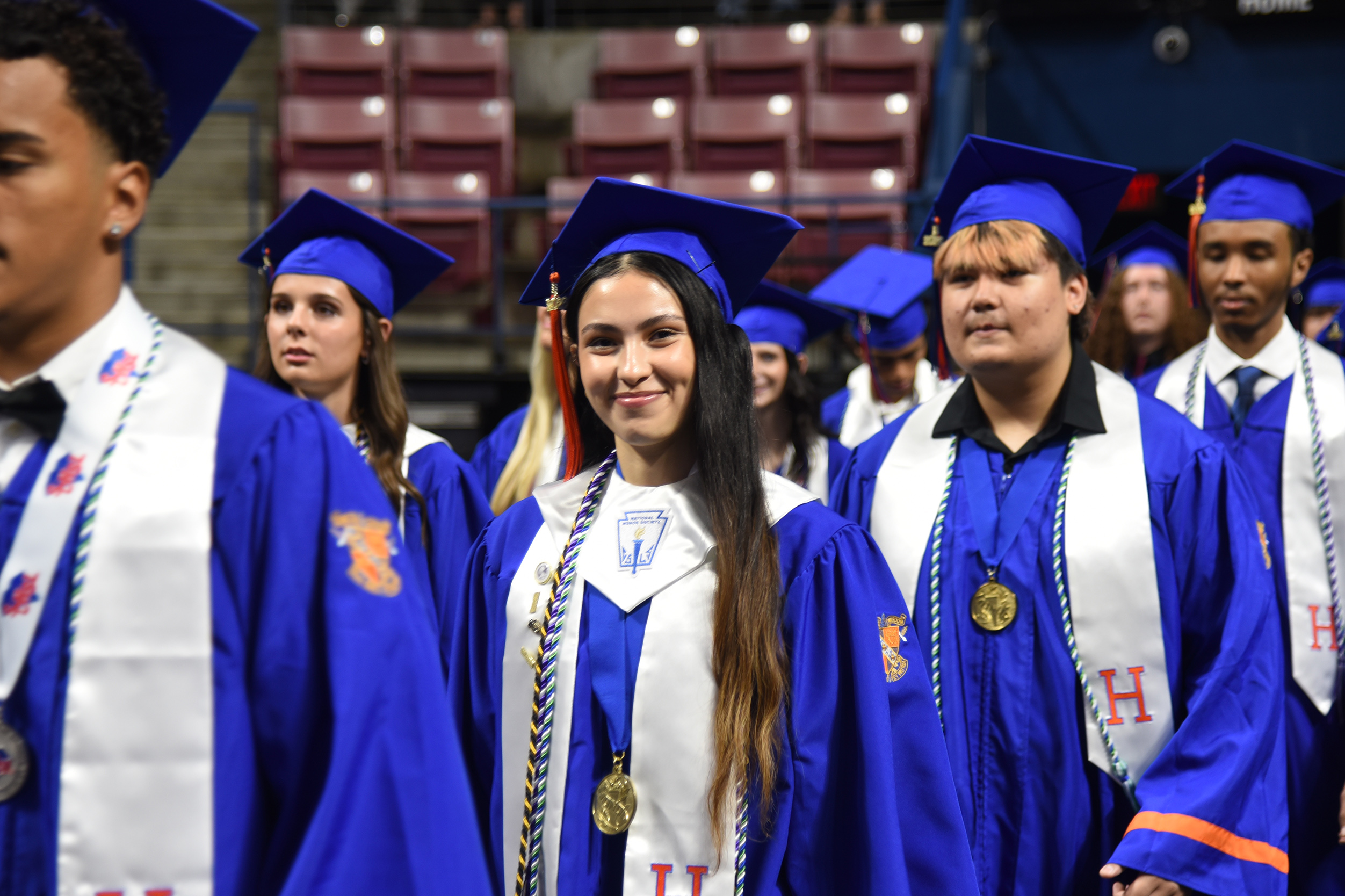 Graduating students in blue gowns and caps stand in a line in an empty auditorium.