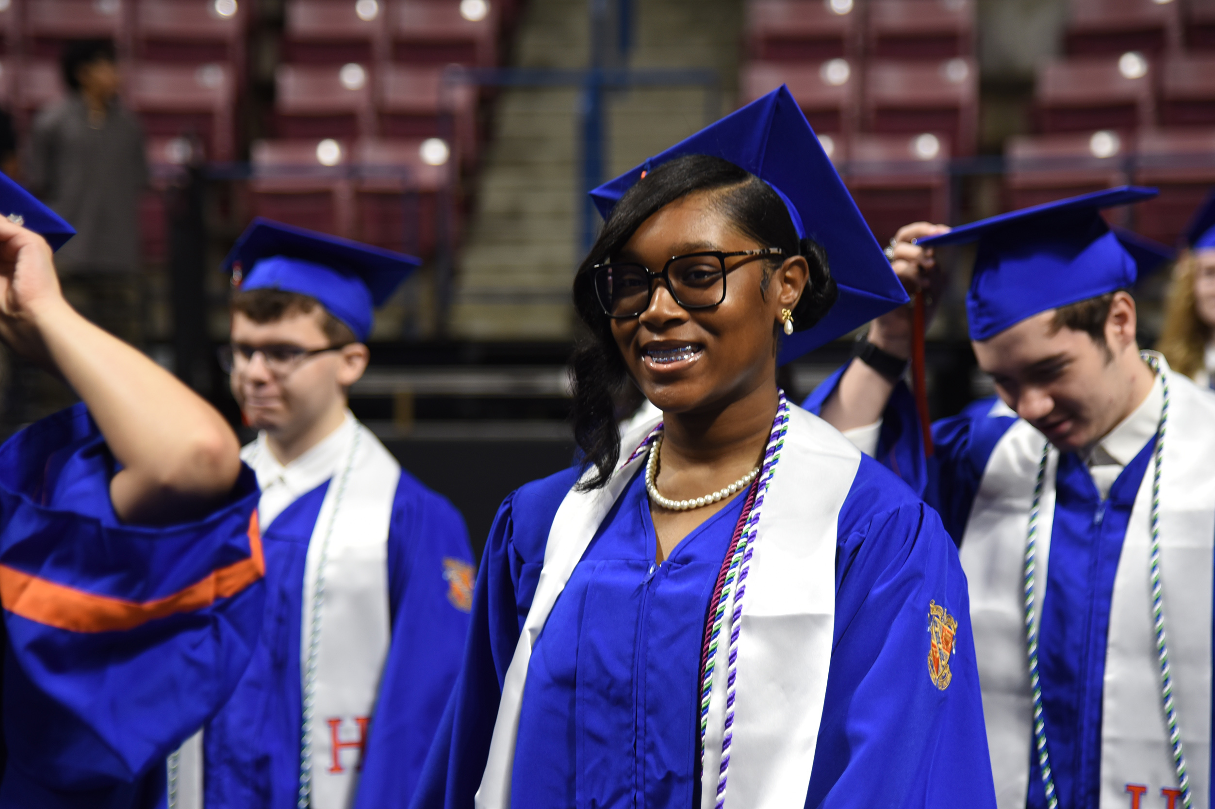 Graduates wearing blue caps and gowns stand in a line. One woman smiles at the camera.