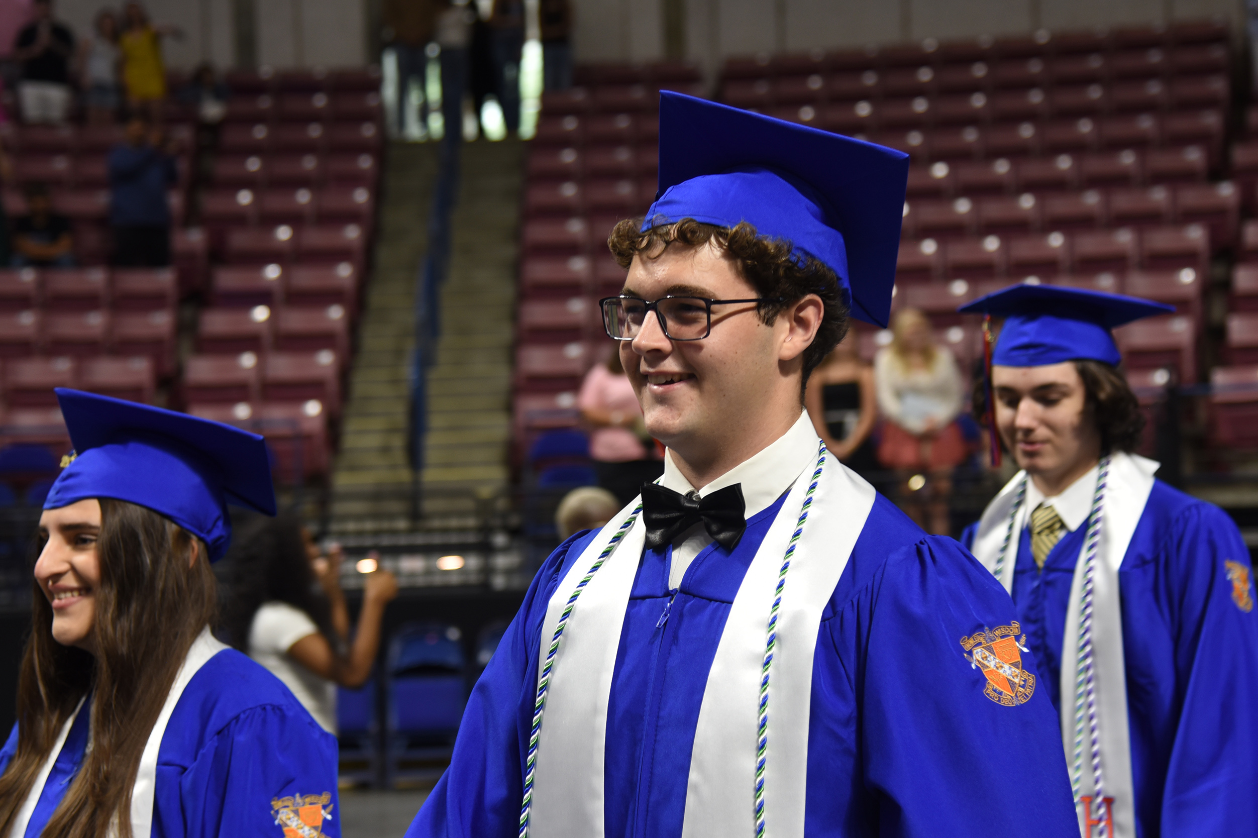 Graduates in blue gowns and caps, with one smiling, stand in an auditorium. Empty seats and staircases in the background.