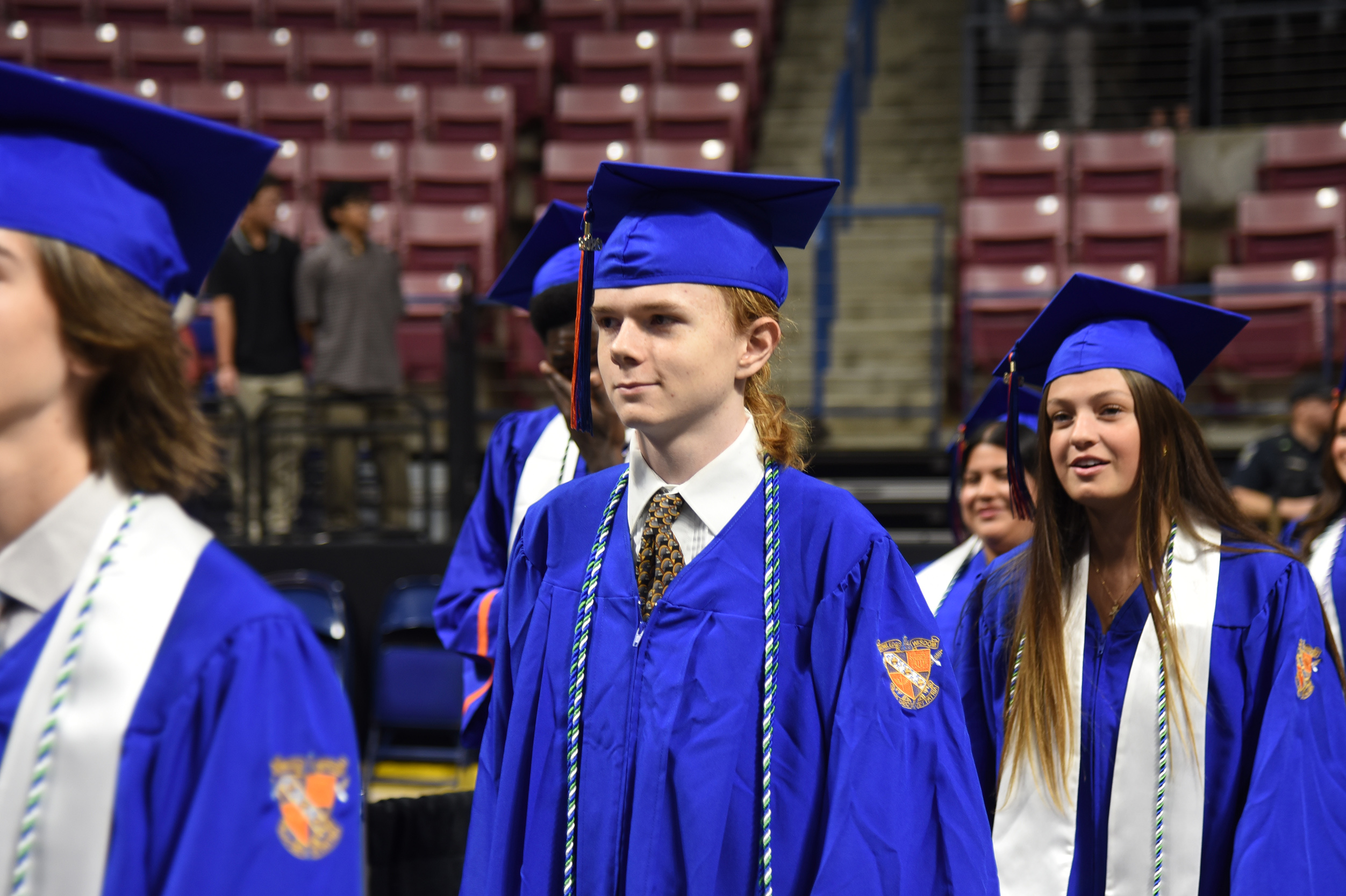 Graduates in blue caps and gowns walk in a stadium, lined with red seats in the background.