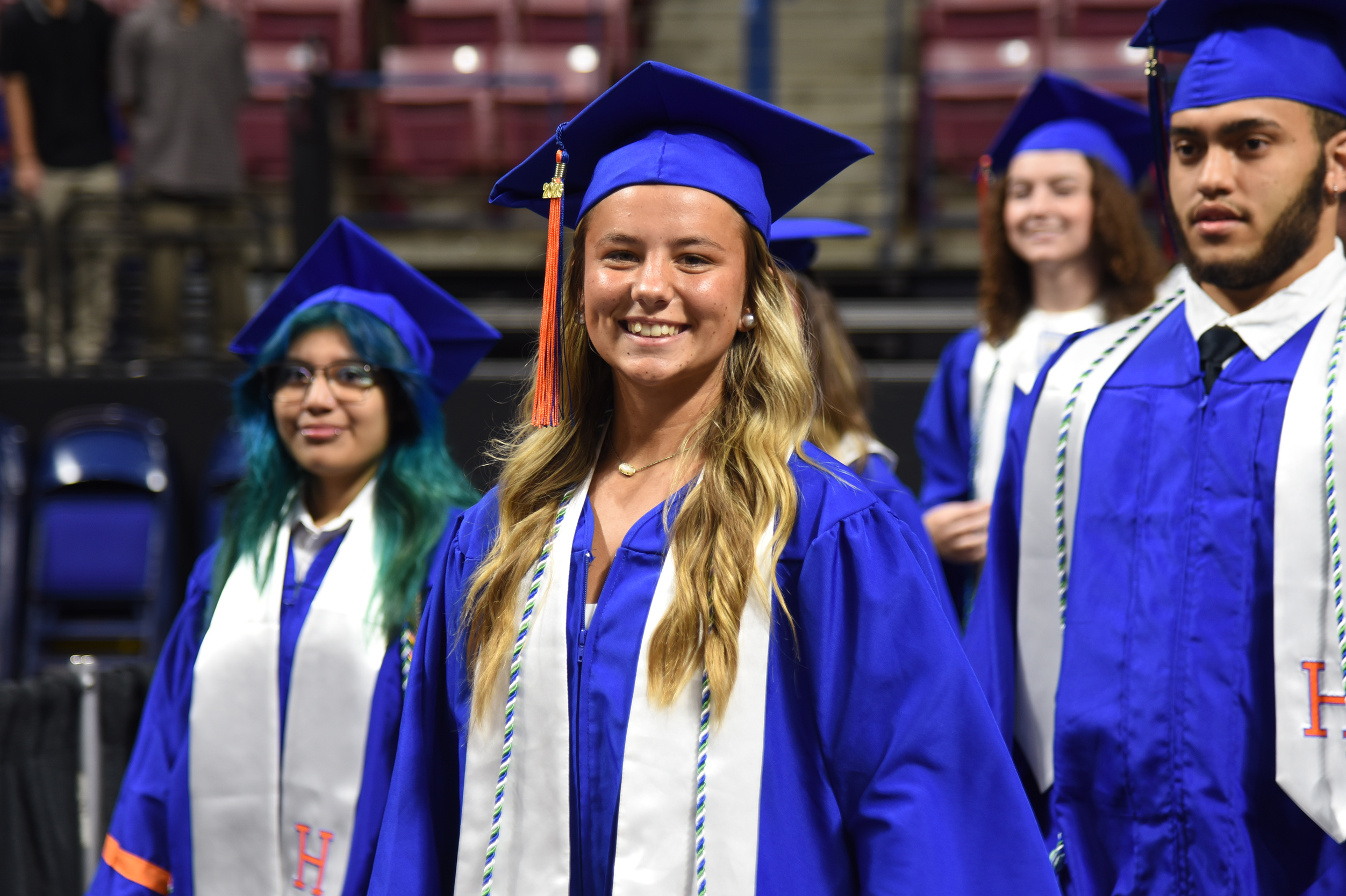 Graduates in blue gowns and caps, walking in an indoor stadium, some wearing sashes.