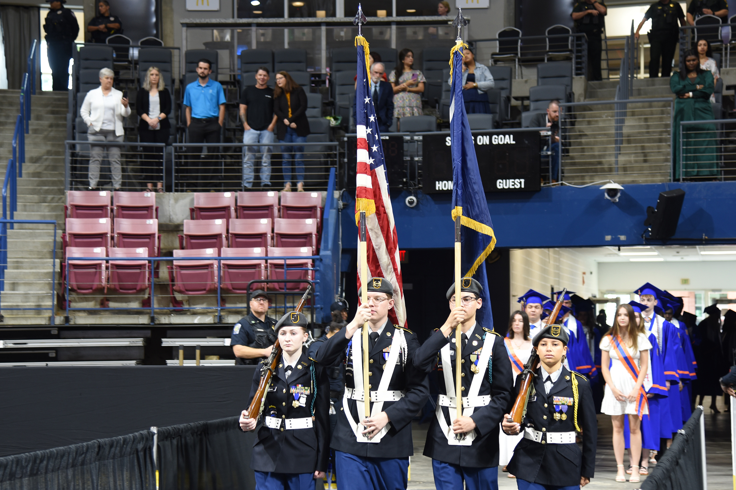 Six individuals in military dress carry flags, including the American flag, in front of a partly filled audience.