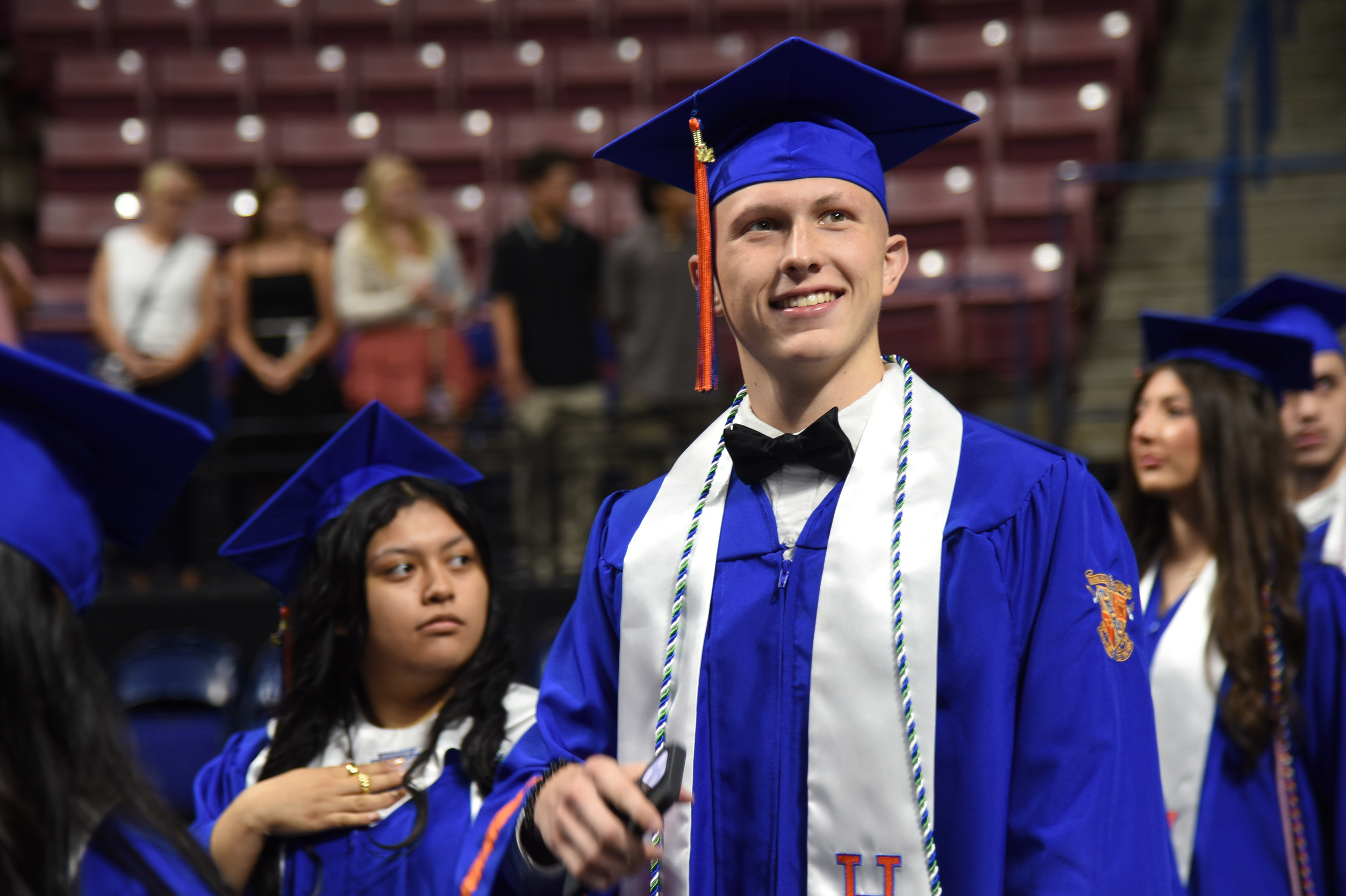A young man in a blue gown and cap stands in a row of people at a ceremony.