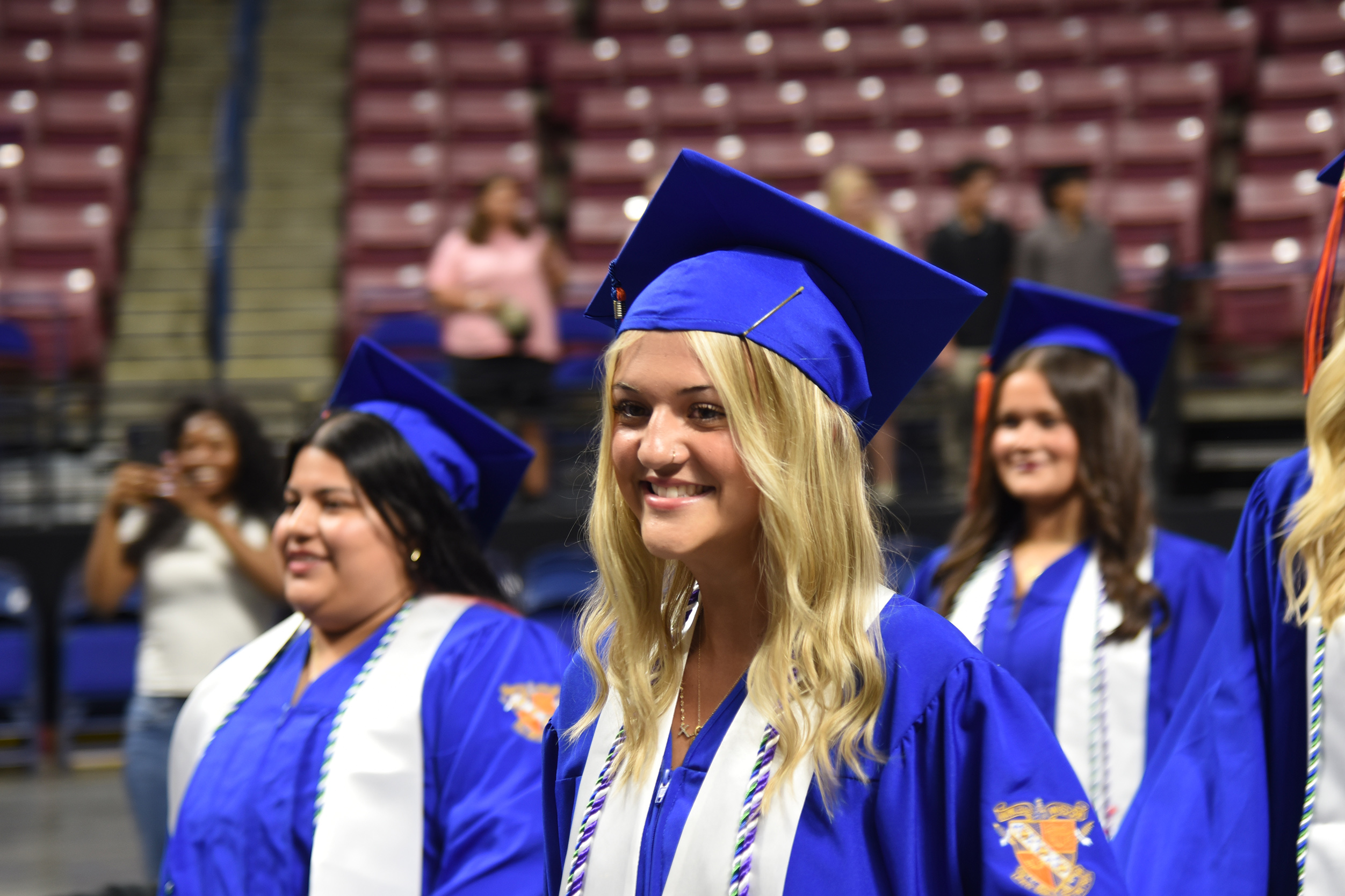 Several women in blue graduation gowns and caps smile at an indoor event; people in the background wear casual clothes.