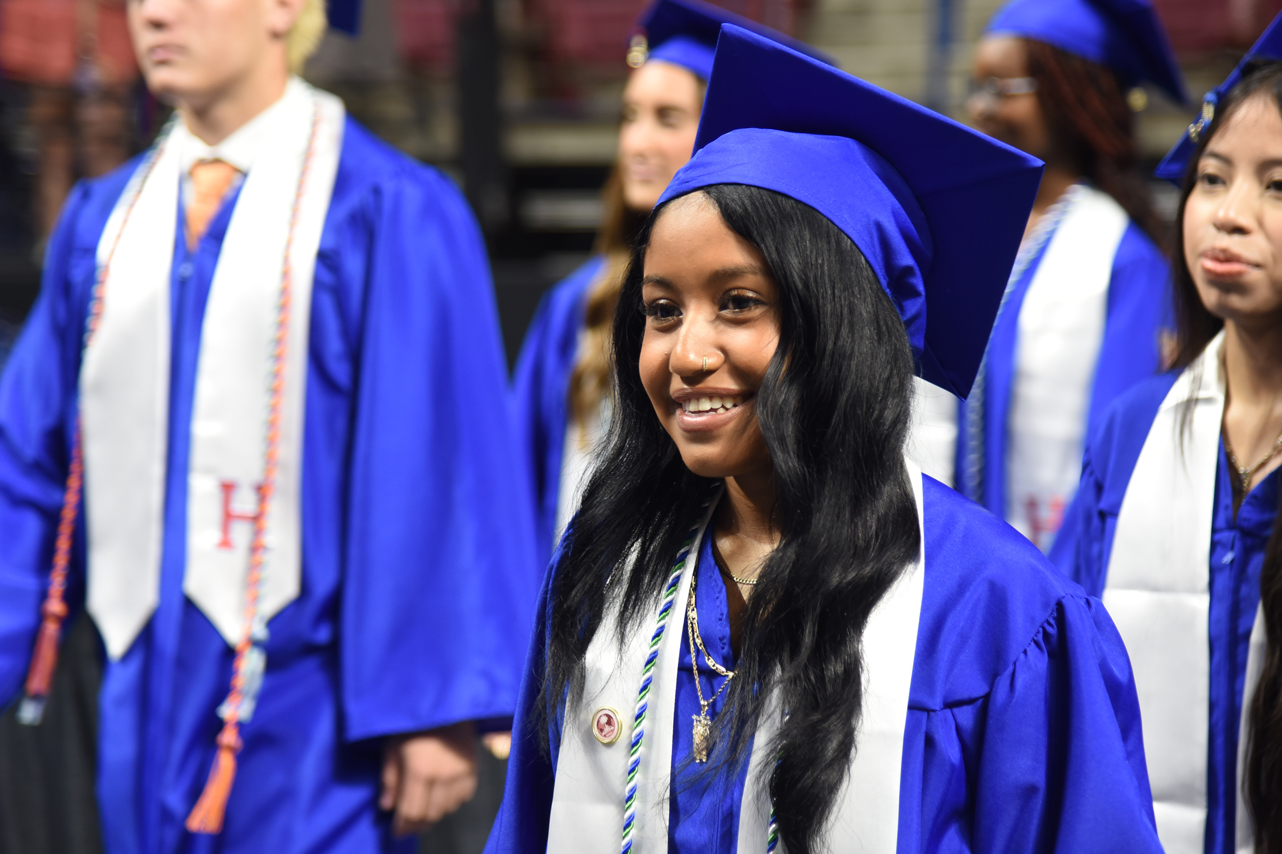 A woman in blue and white graduation attire stands among others in a line, wearing a blue cap and smiling.