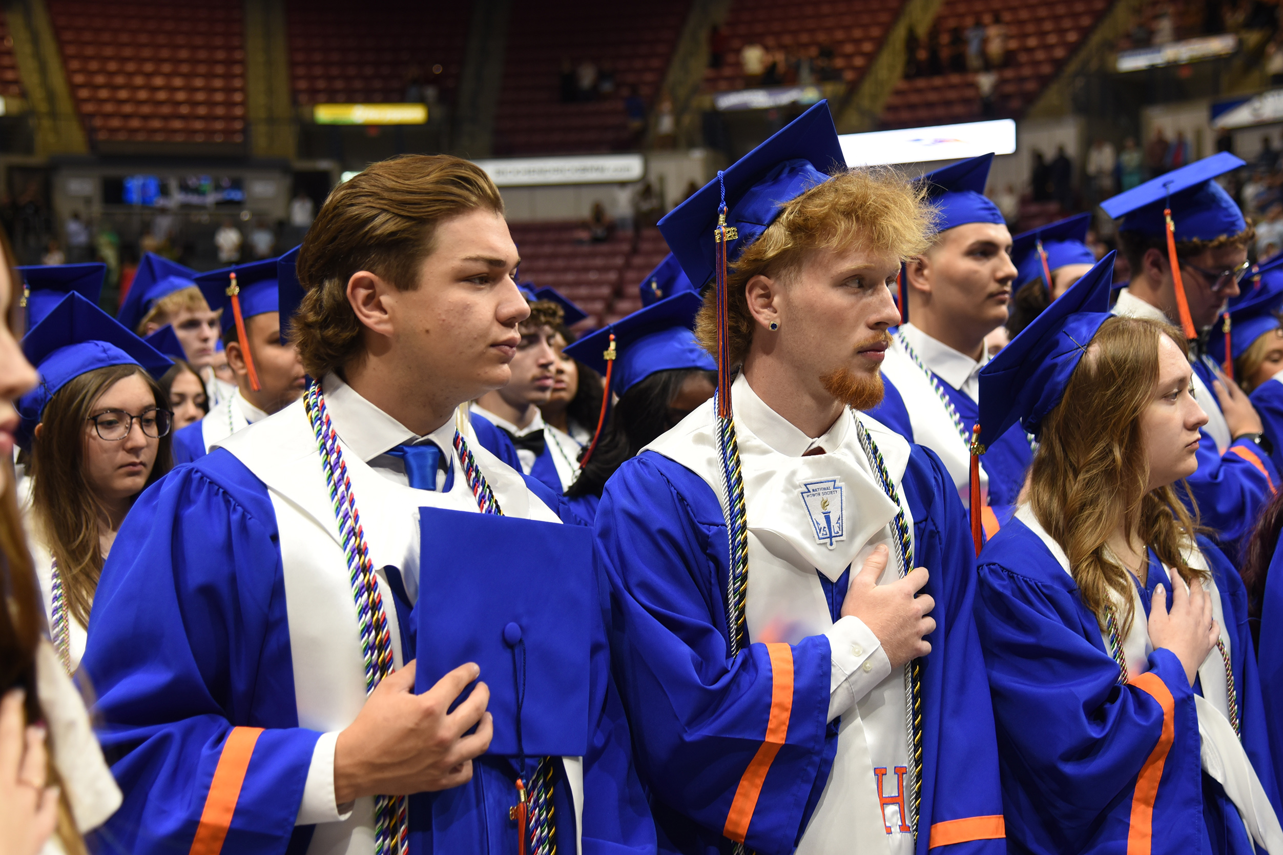 A row of students in blue and white graduation gowns, hats, and scarves stand in a stadium.