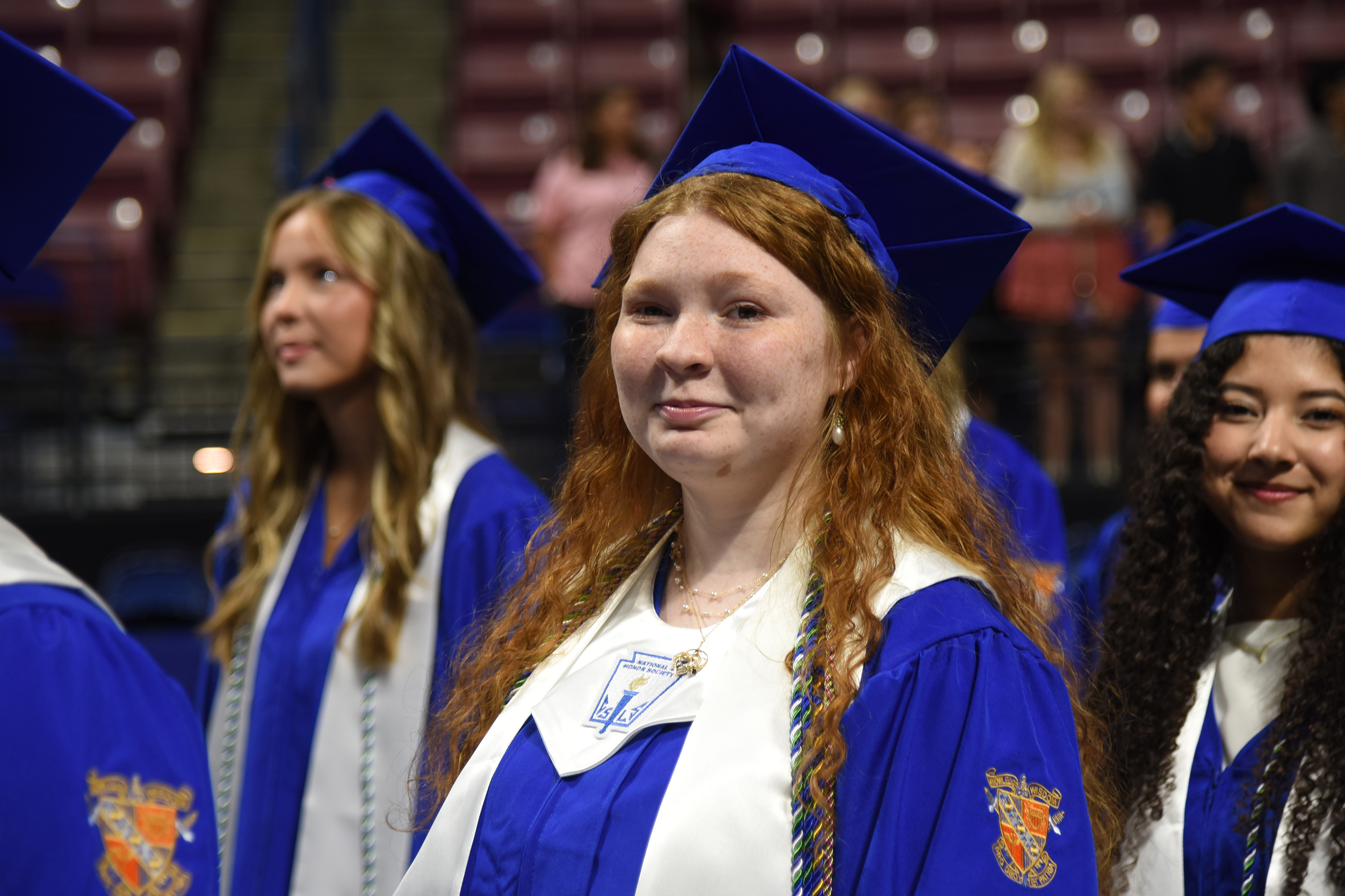 Four women in blue and white graduation gowns and caps at an event in a large venue.