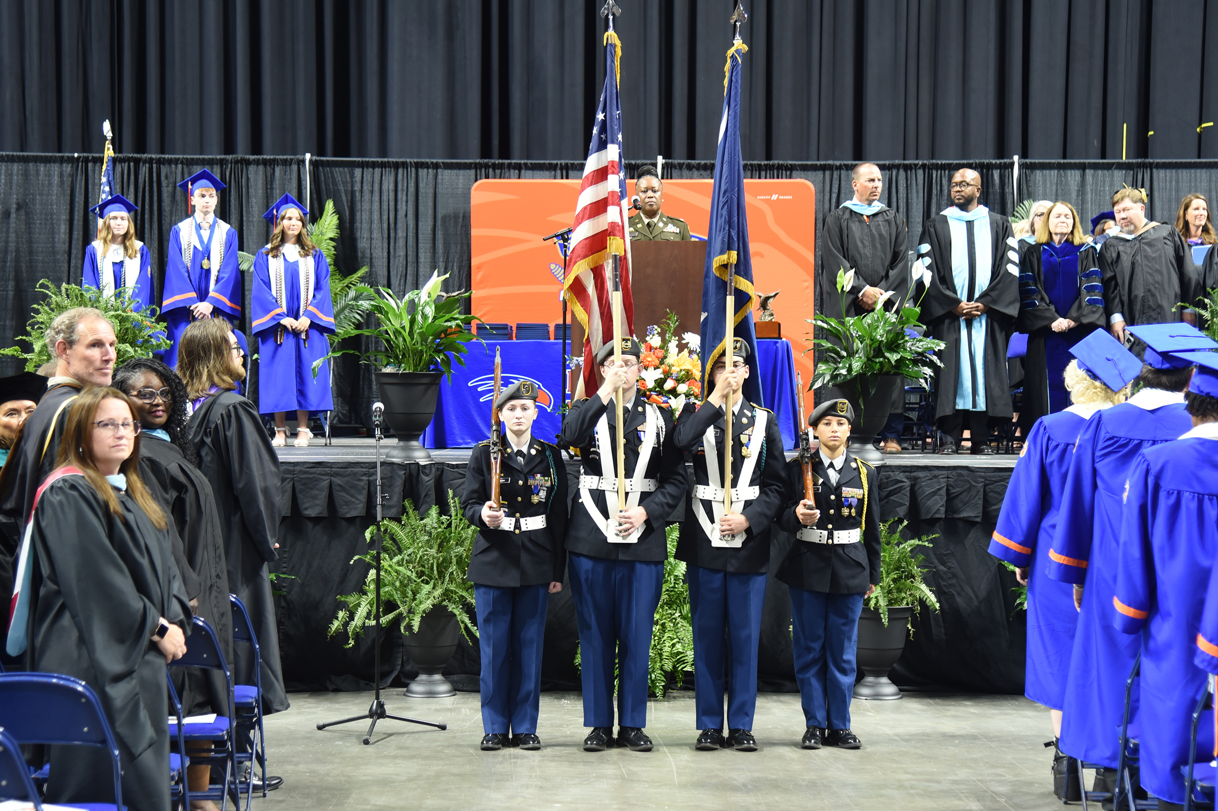 A group of people in blue gowns and hats stand on a stage, with a podium and flags.