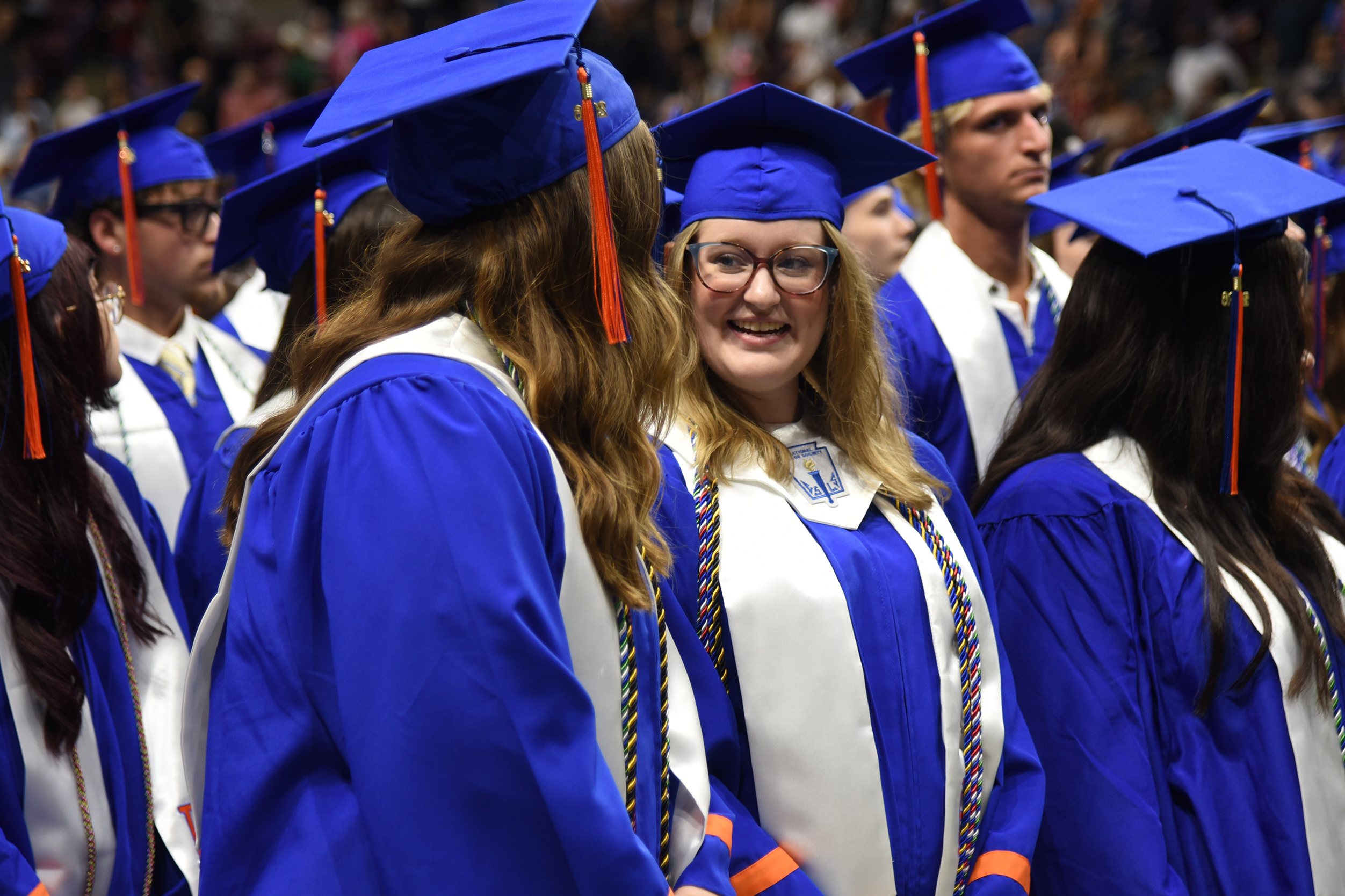 Several students in blue caps and gowns stand in a row at an event, some wearing glasses.