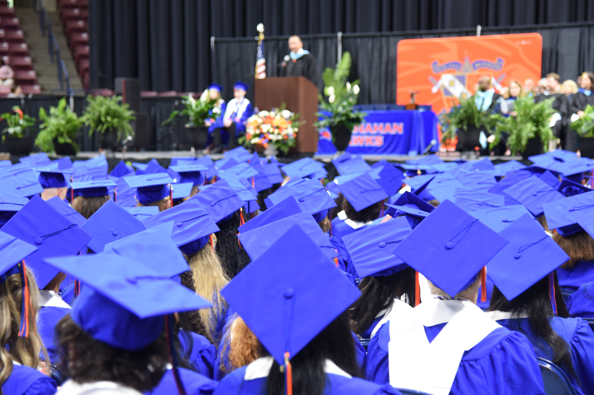 Graduates in blue caps and gowns sit in rows, looking up at the stage where a person is speaking.