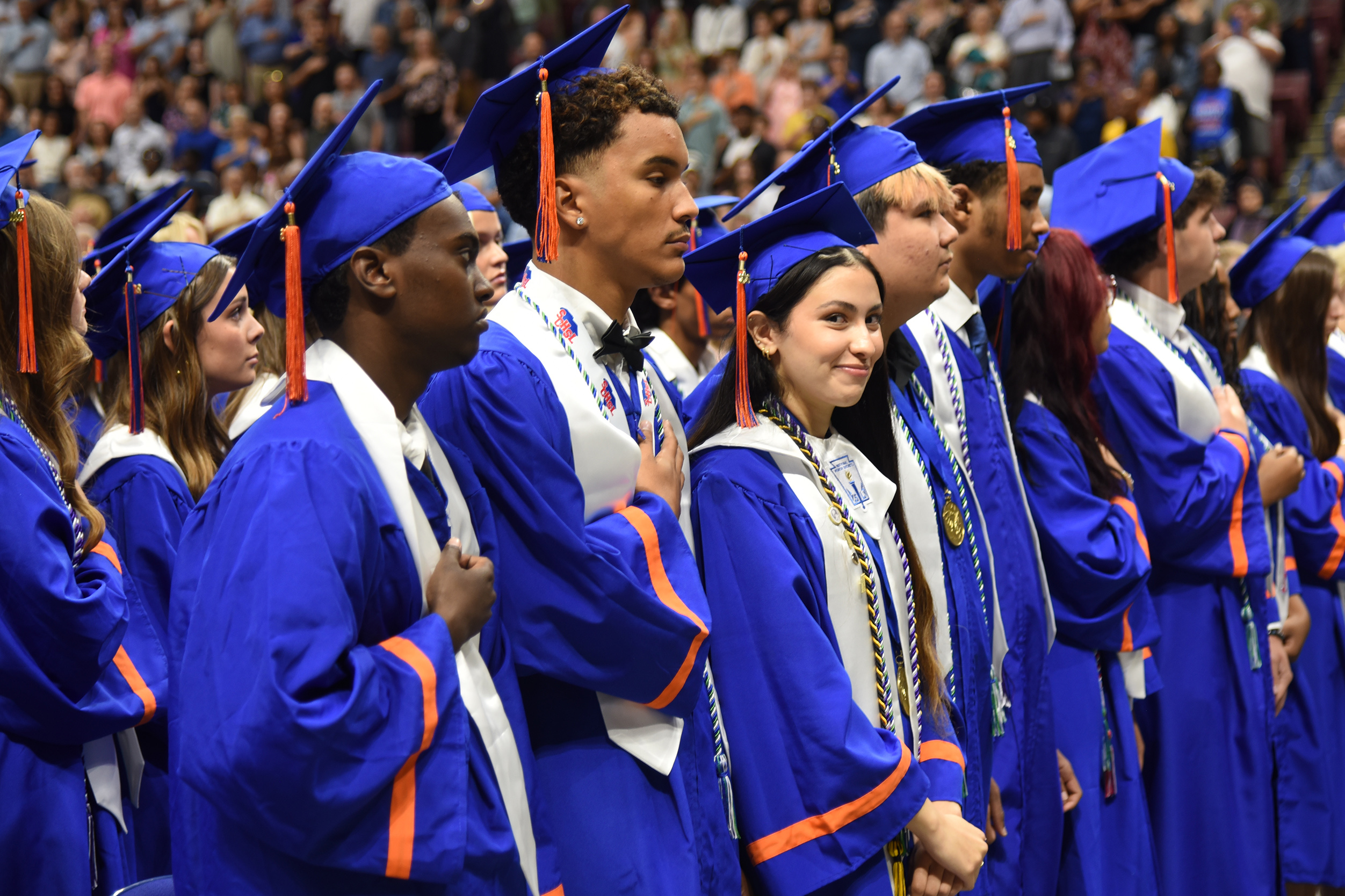 People in blue gowns with orange trim and tasseled hats line up. They face the audience at a school event.
