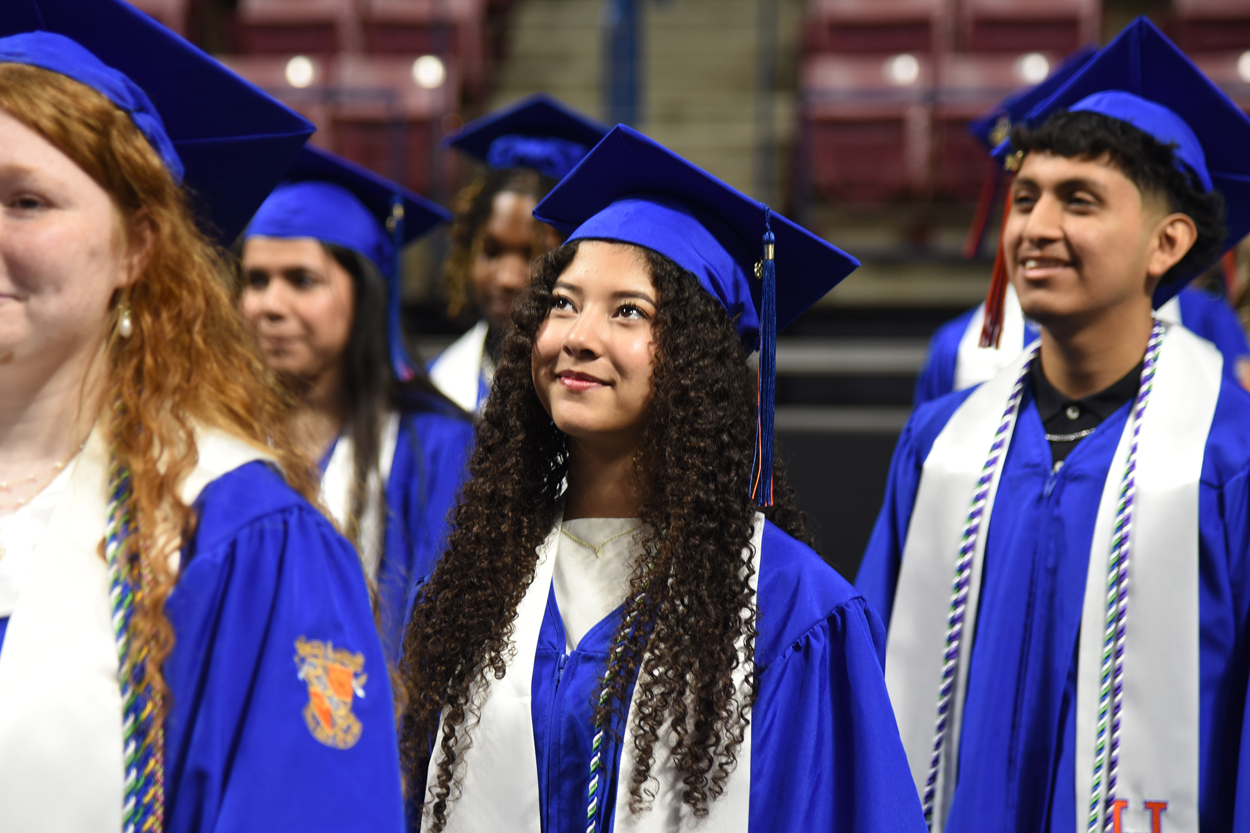 Graduates in blue and white caps and gowns stand in a row. A woman with curly hair looks to the right.