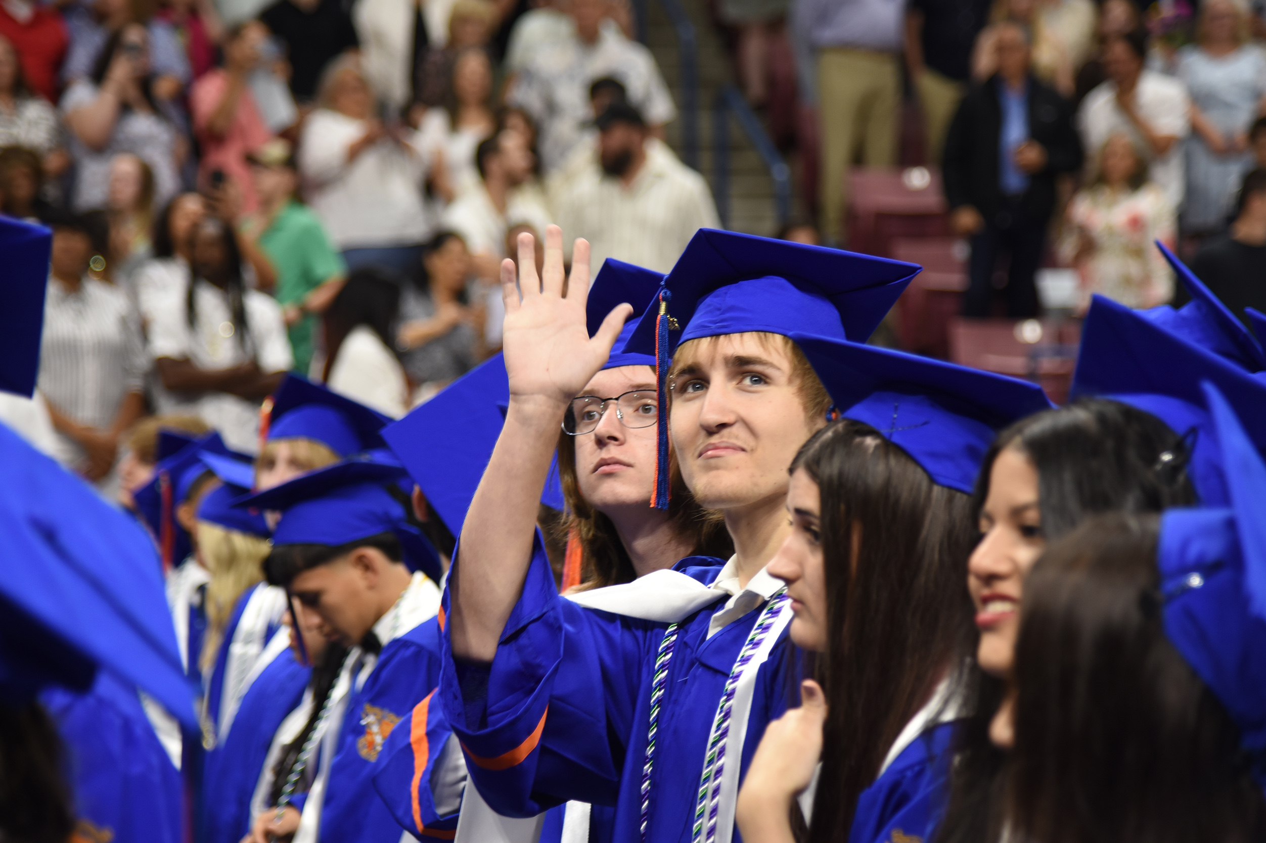 Graduates in blue gowns and caps, one raising a hand, stand among an audience in an auditorium.