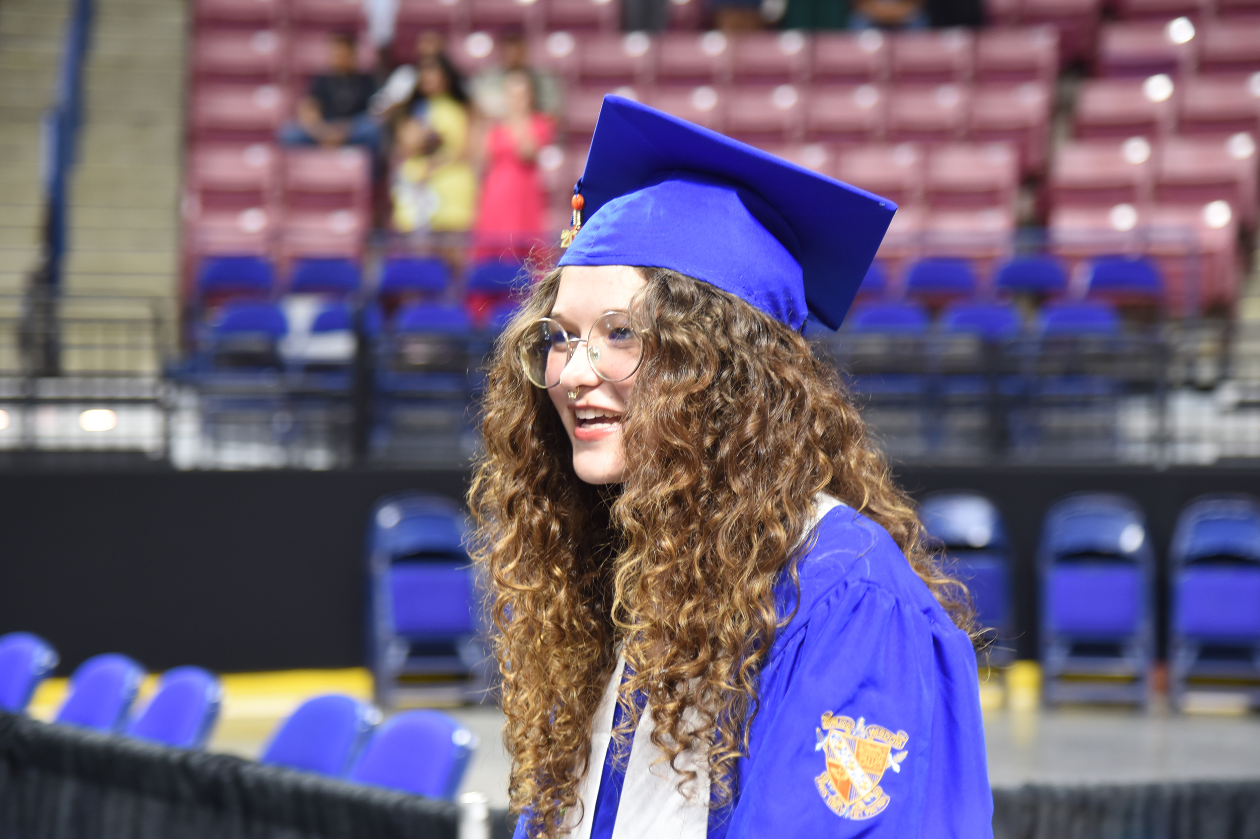 A person with curly hair in a blue gown and hat stands in a stadium with empty blue chairs.