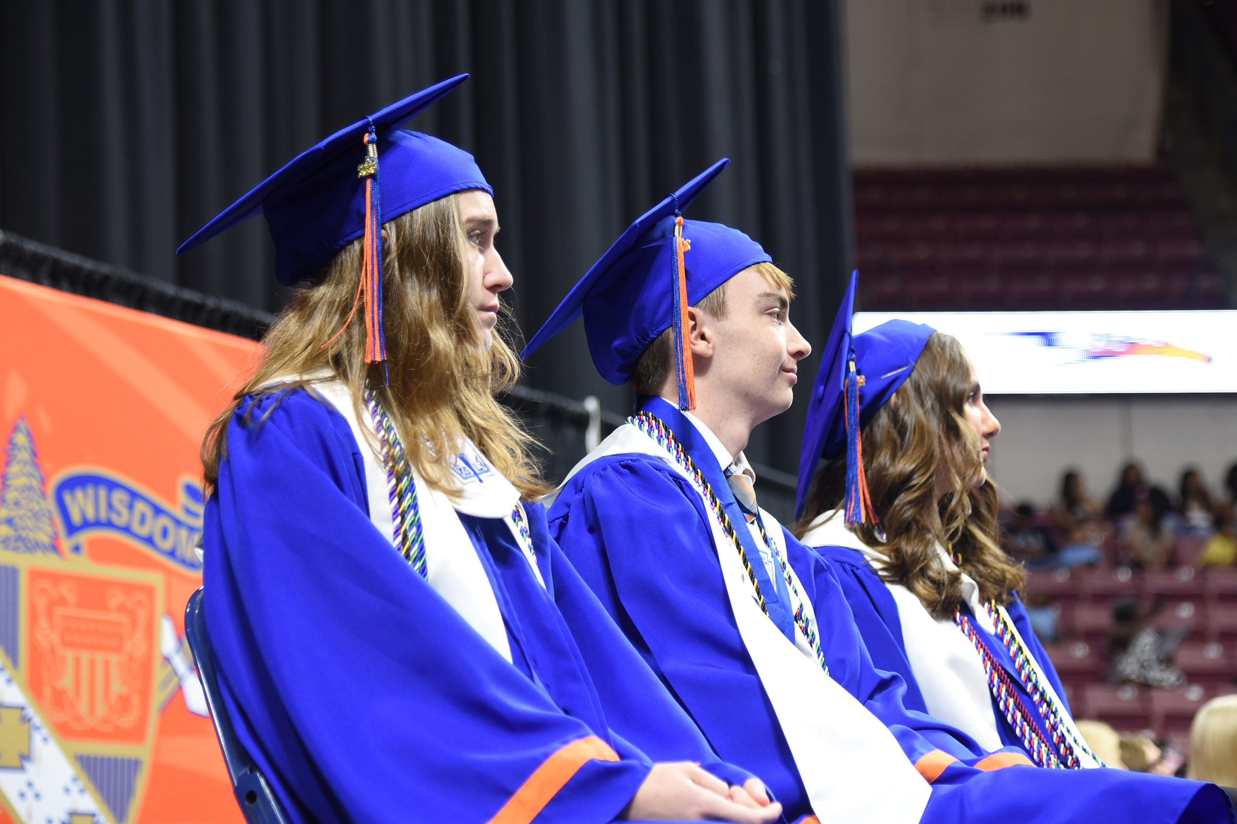 Graduates in blue gowns and caps sit in an auditorium. An orange banner with a logo and text is behind them.