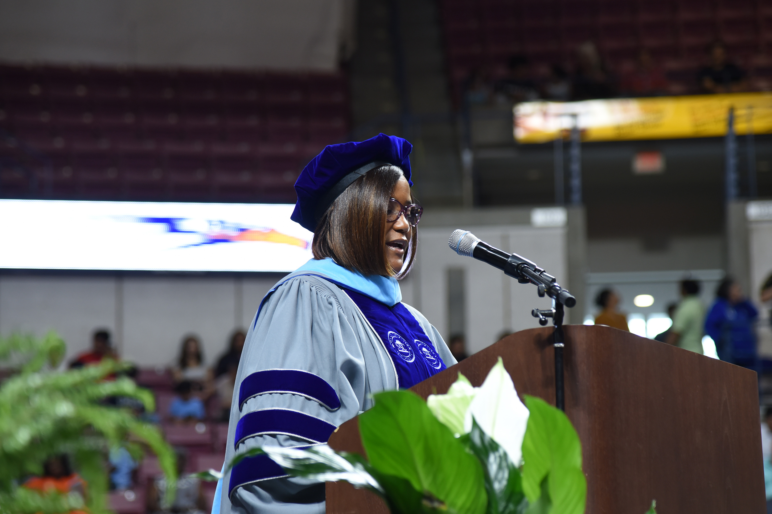 A woman in a cap and gown speaks at a podium with a microphone. An audience sits in bleachers behind her.