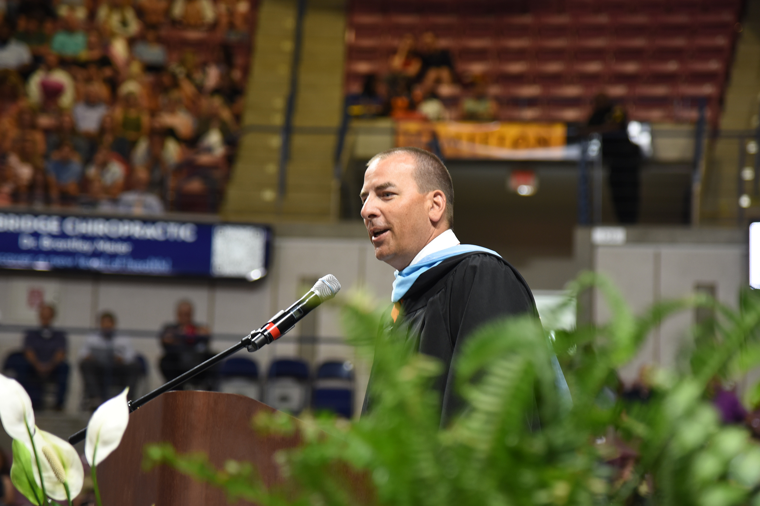 A man wearing a graduation gown speaks into a microphone at an event with plants and an audience.