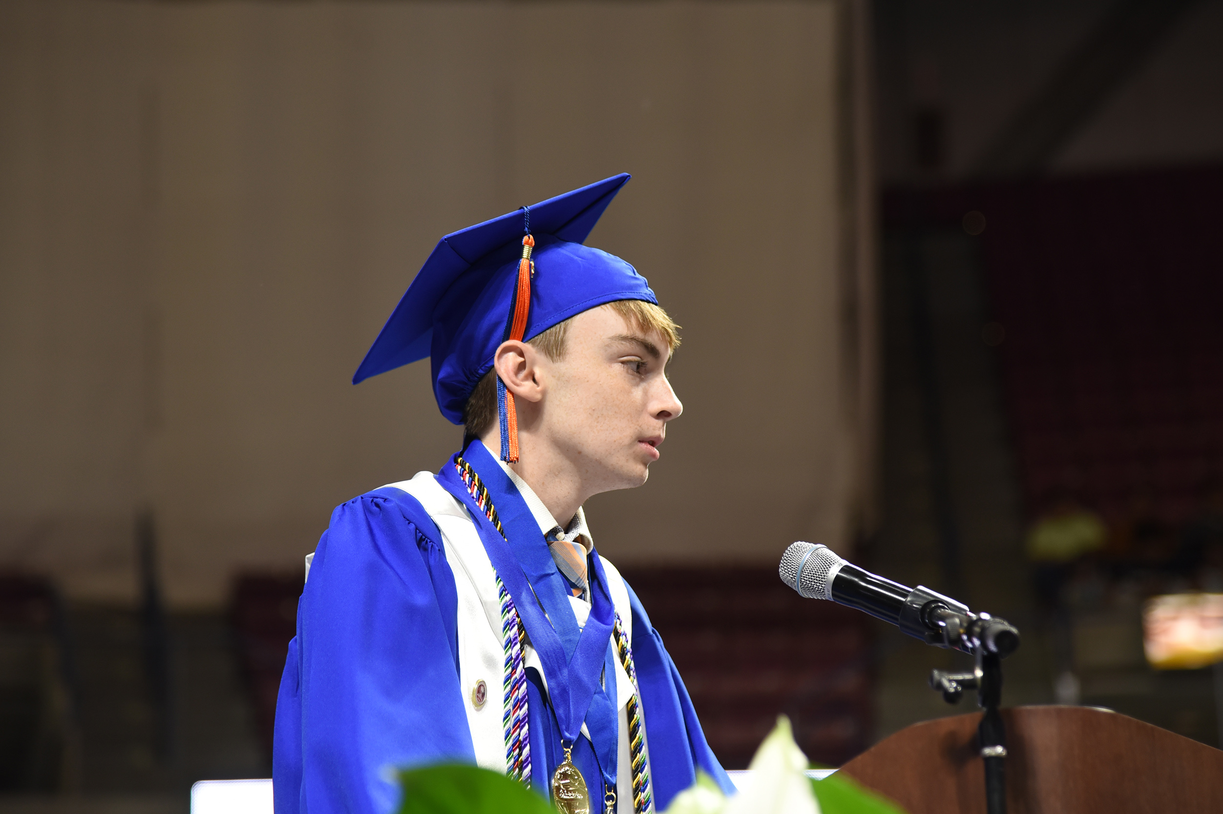 A male in a blue cap and gown stands at a podium, facing forward, with a microphone before him.