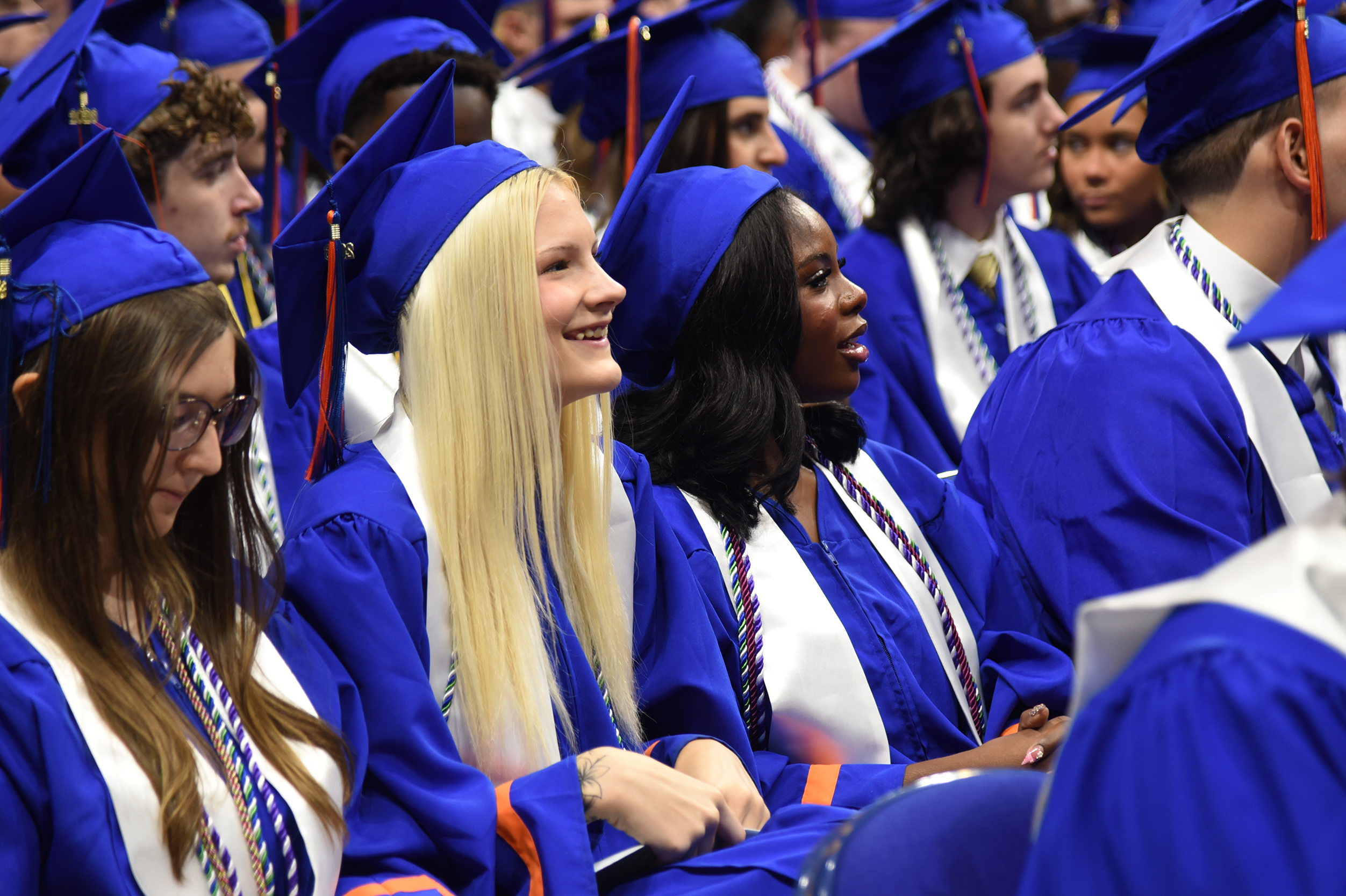 A crowd of students in blue and white graduation gowns and caps, some smiling and looking ahead.