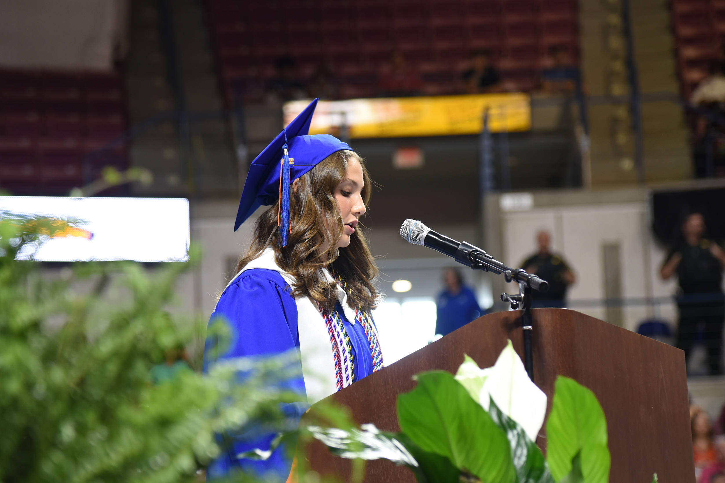A woman in a blue gown and hat speaks into a microphone at a podium in a venue.