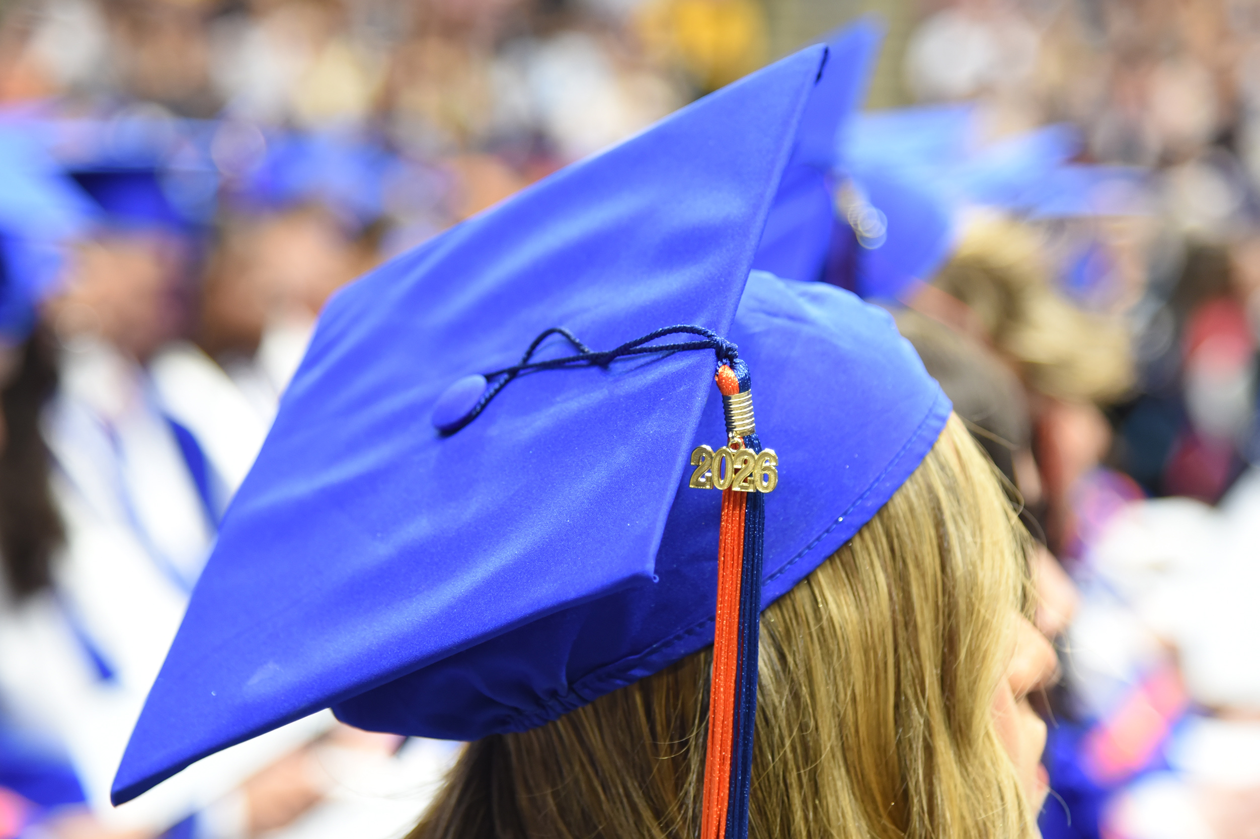A close-up of a blue graduation cap with tassel and the year 2026 in gold letters, worn by a person with blonde hair.