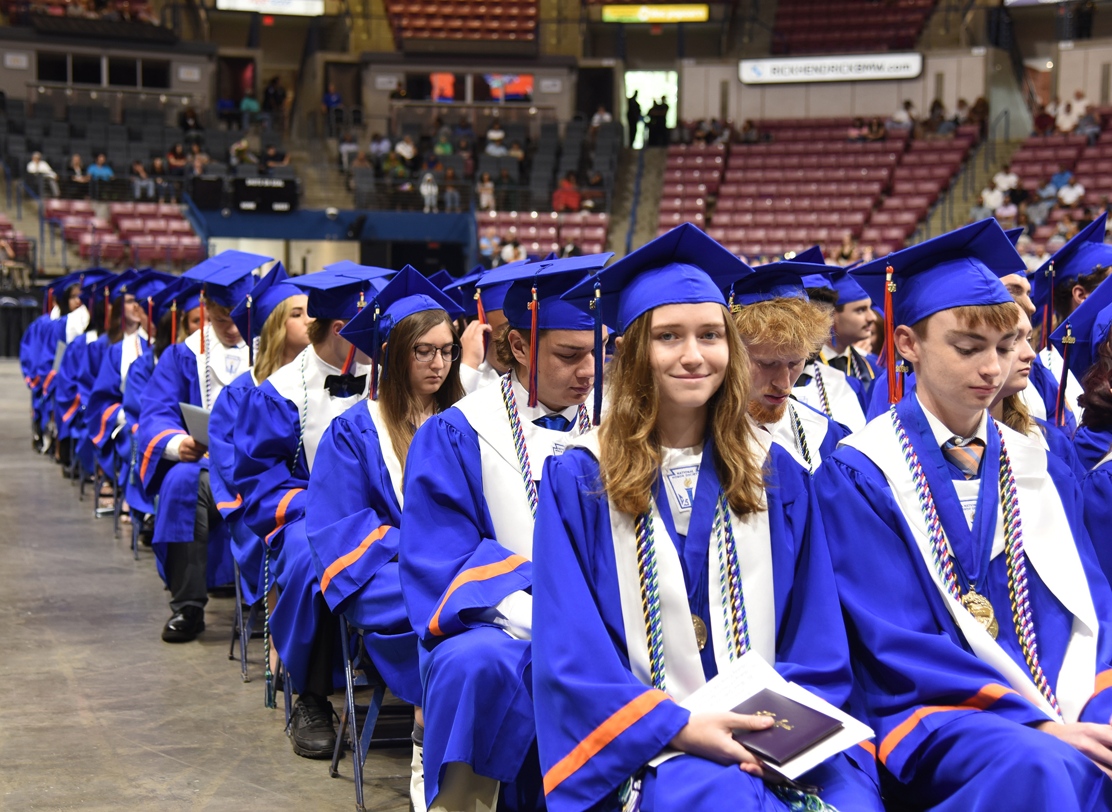 Graduates in blue gowns and caps sit in an arena, some looking forward and others down at a book.