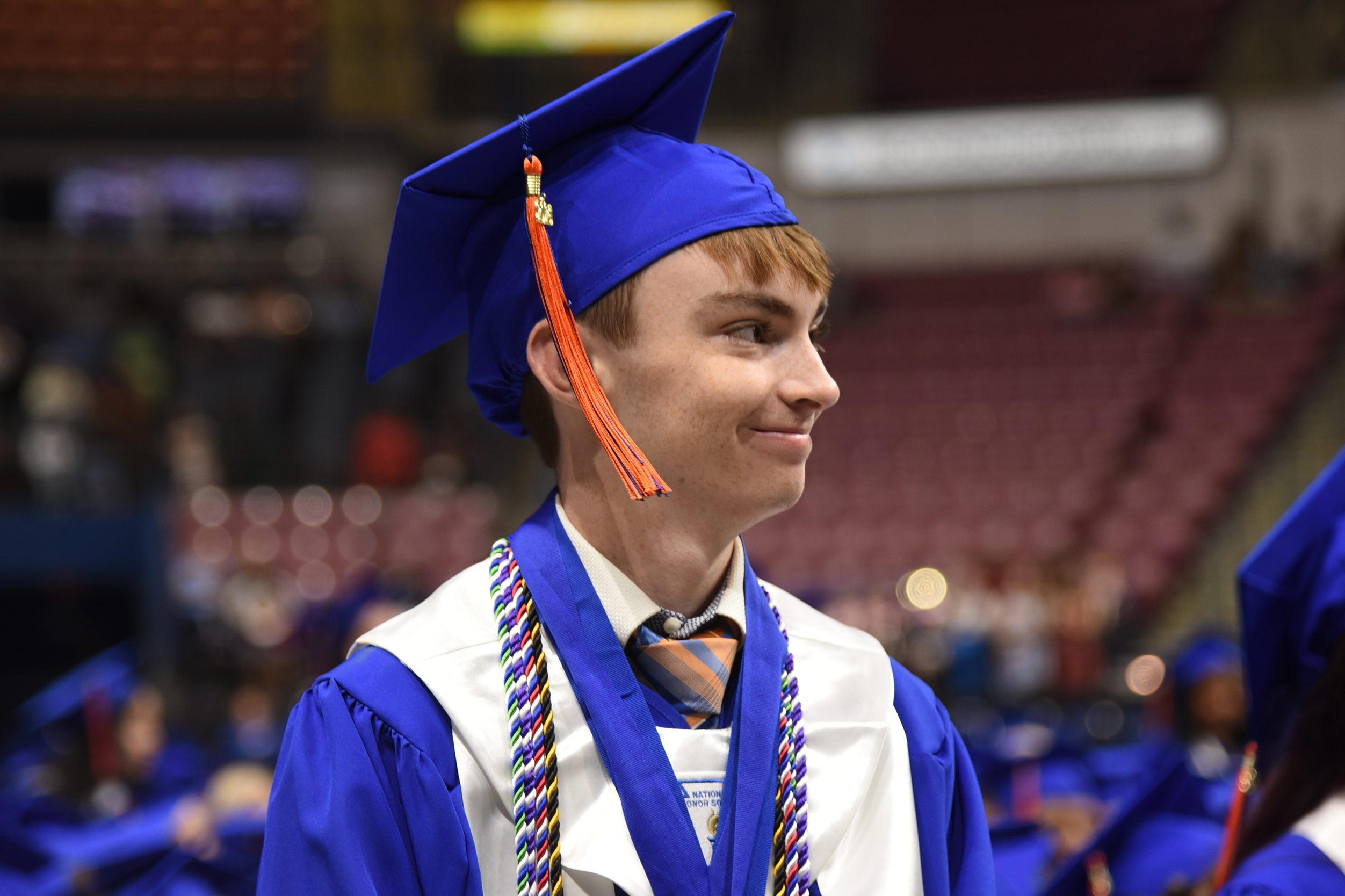 A young person in a blue cap and gown stands in an auditorium, smiling. Behind them, empty red chairs are visible.