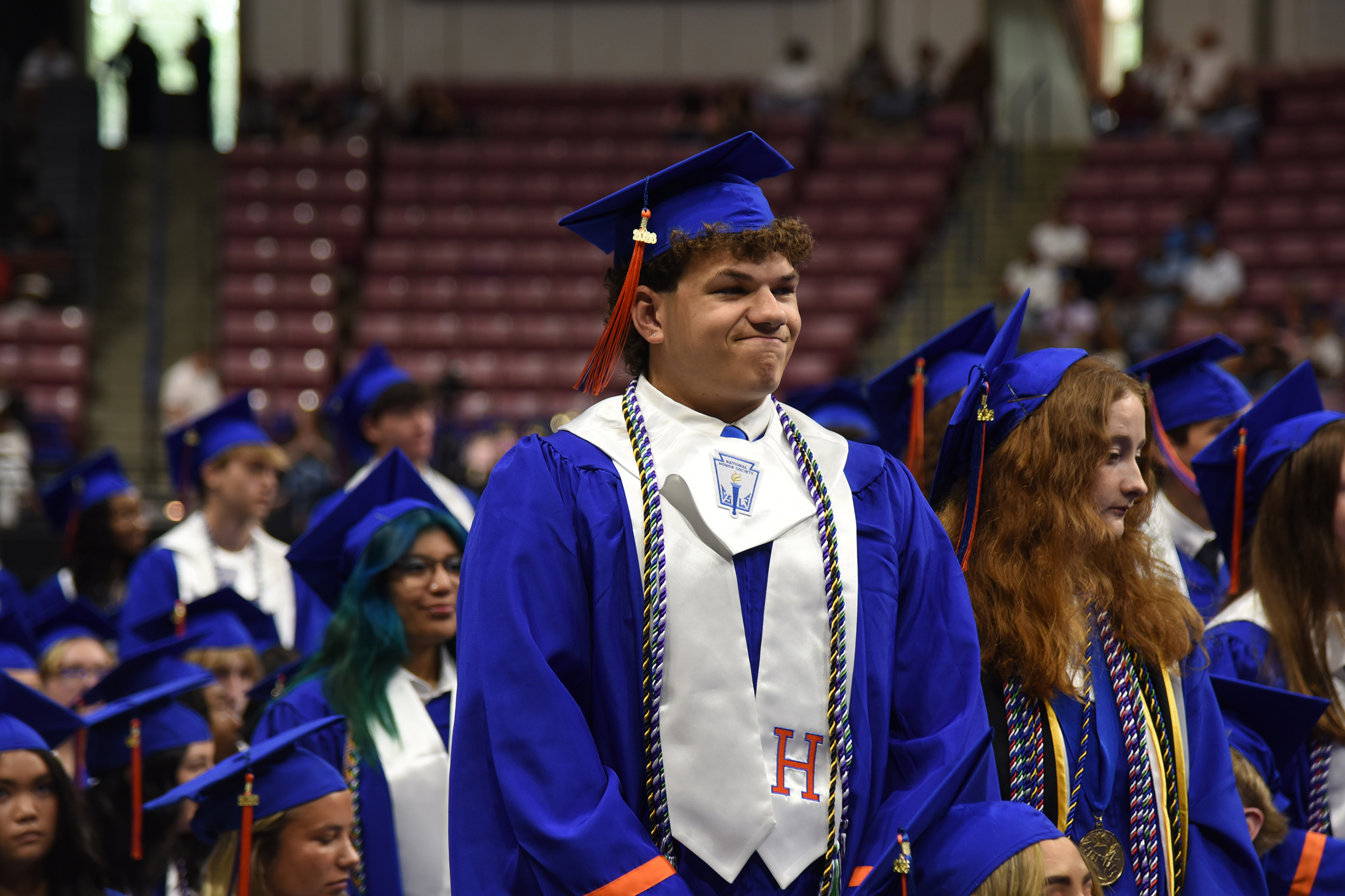 A group of people wearing graduation gowns and caps stand in a stadium with red seats.