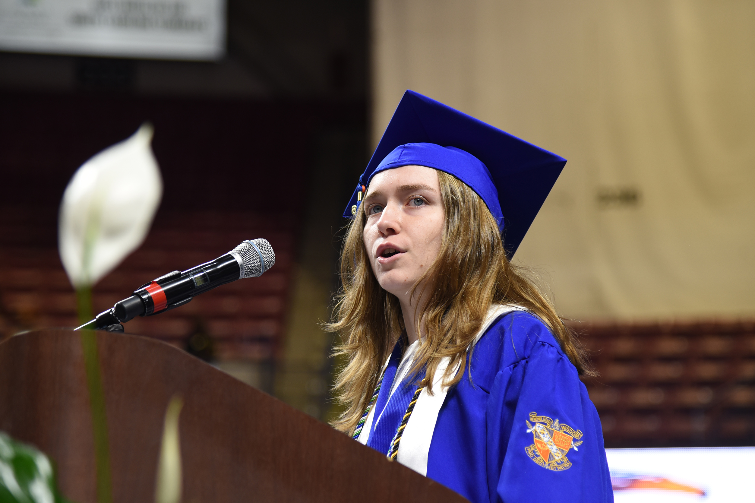 A woman in a graduation gown speaks into a microphone at a podium, with a white flower behind her.