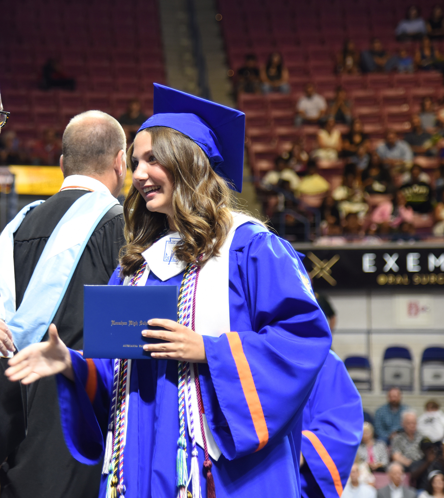 A woman in a blue graduation gown holds a diploma in her right hand. She smiles and stands in front of a crowd.