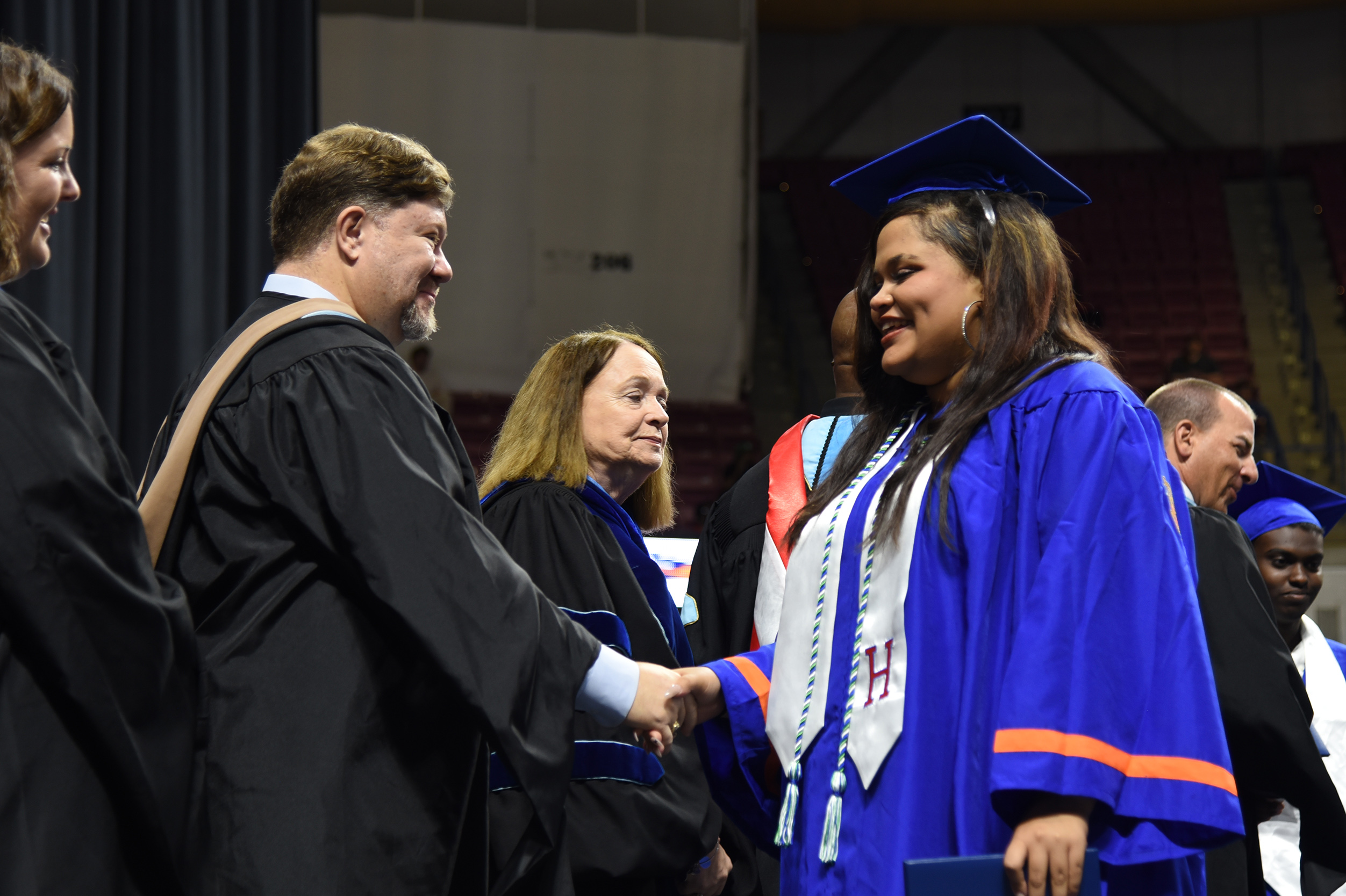A man in a black gown shakes hands with a woman in a blue gown, both smiling. Behind them, several people wear graduation attire and stand in a large room.