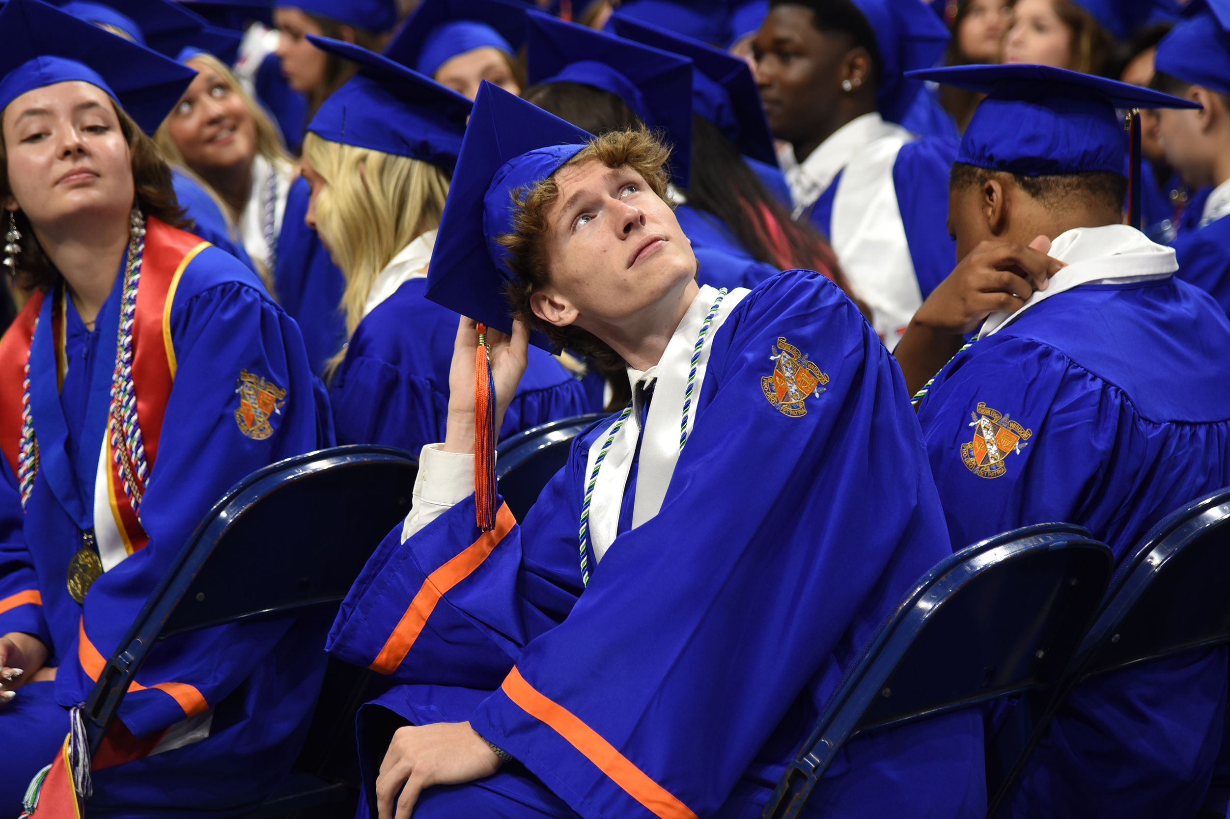 A group of students in blue and orange gowns and caps are sitting in chairs at a graduation ceremony.