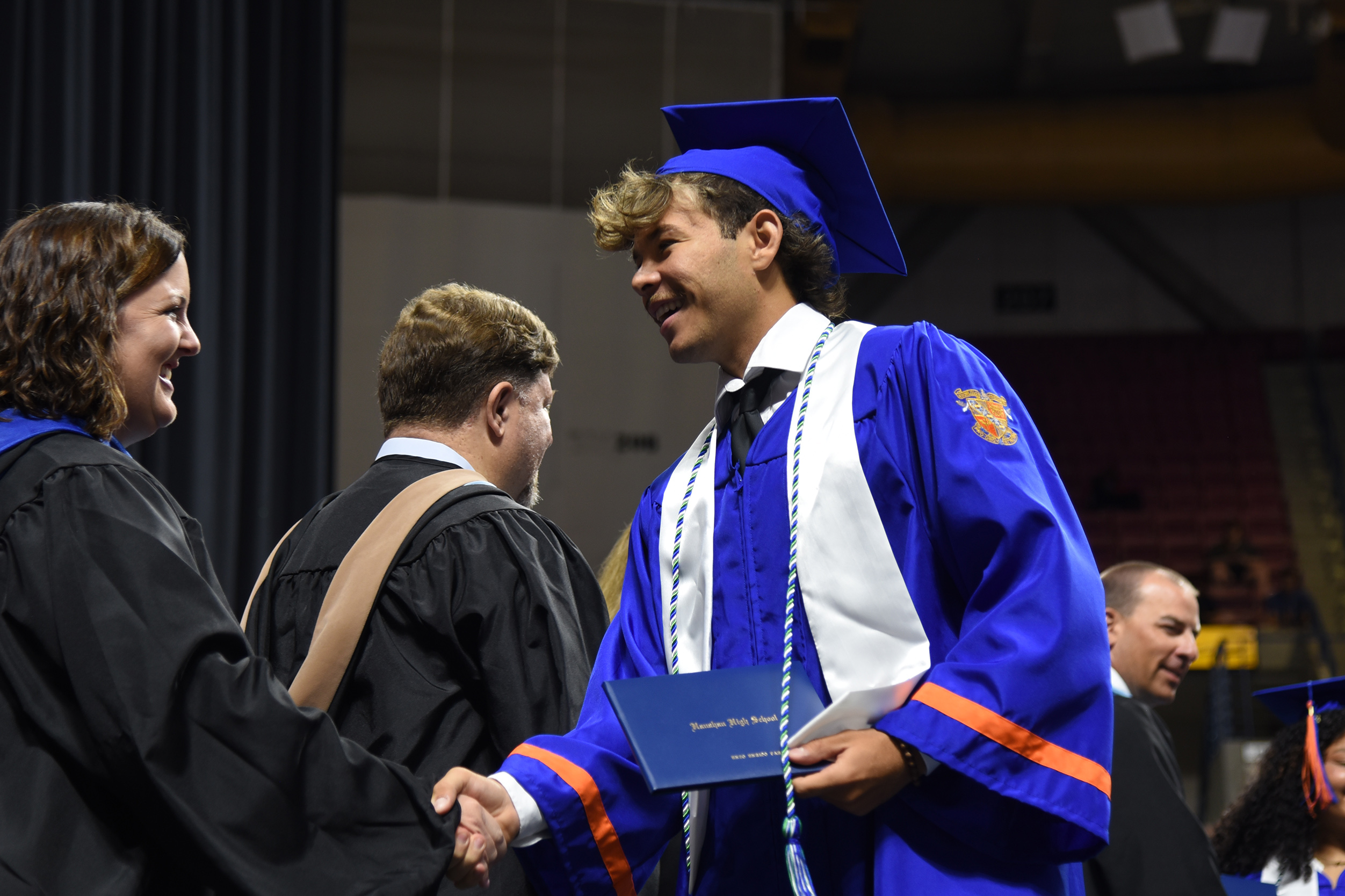 A person in a blue gown shakes hands with a woman in a black gown. They are at a graduation ceremony.