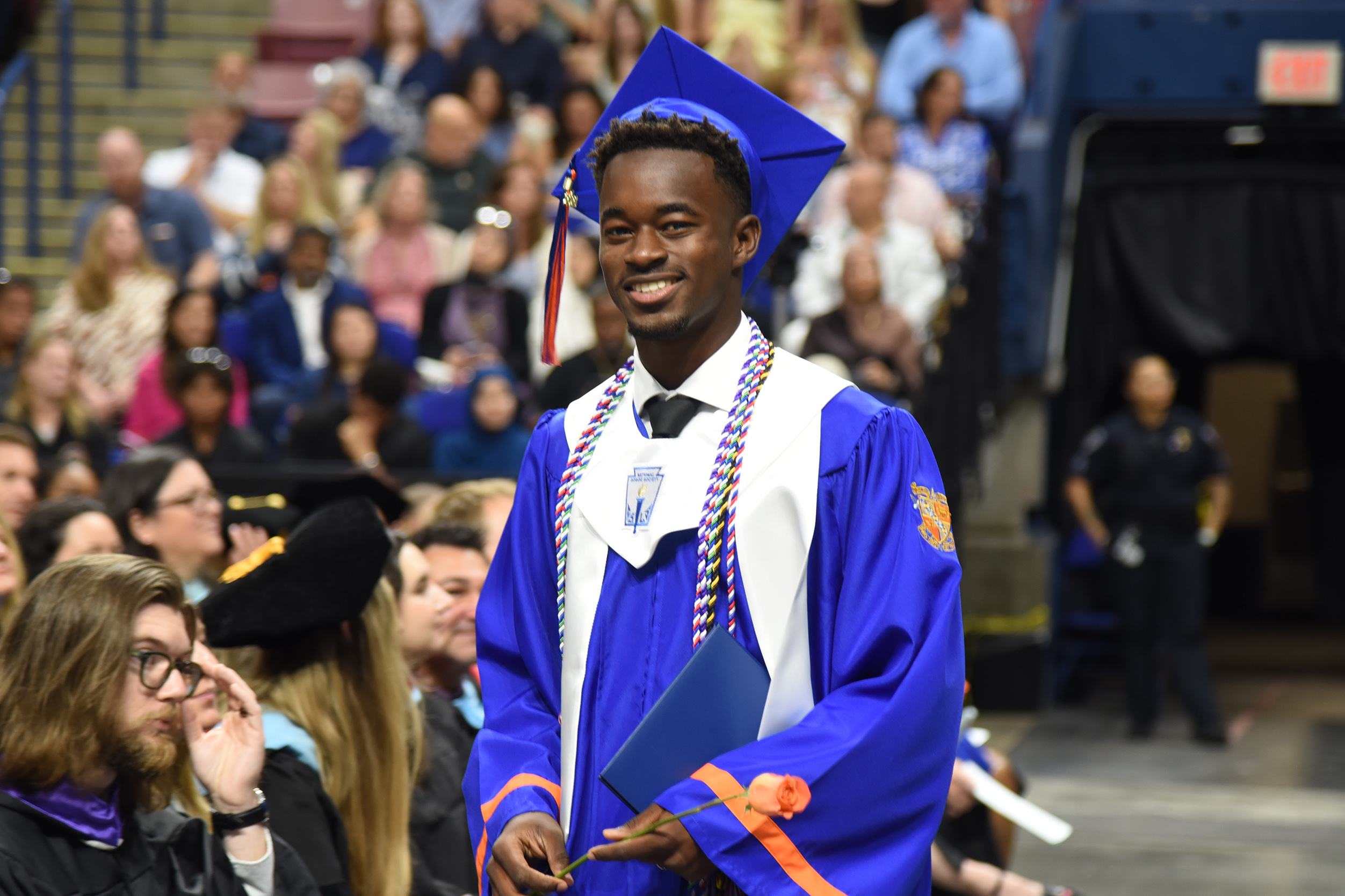 A male graduate stands in front of an audience in a blue gown, smiling. Many people are seated in the background.