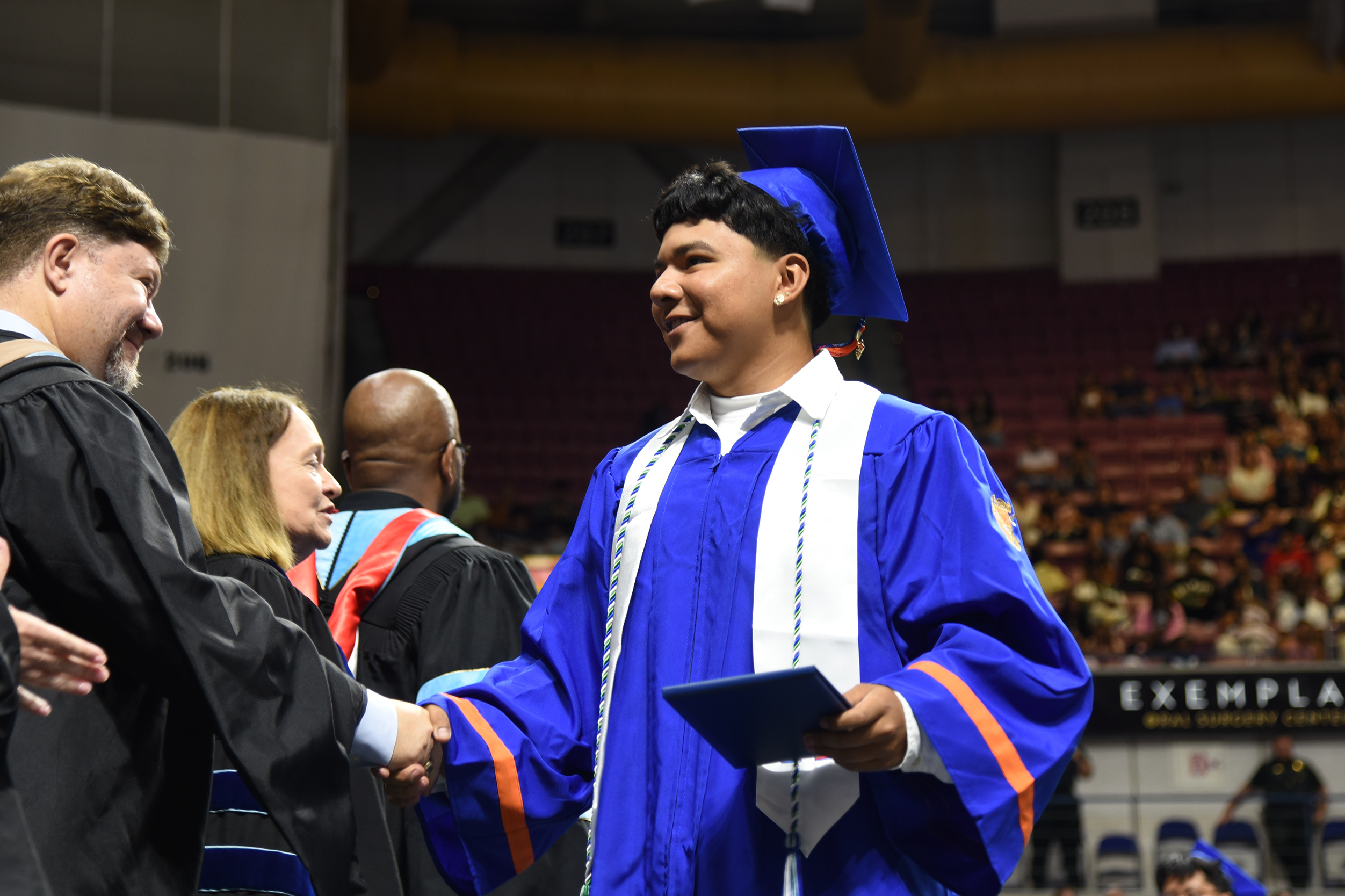 A graduate in a blue gown shakes hands with a faculty member. Audience members sit in the background.