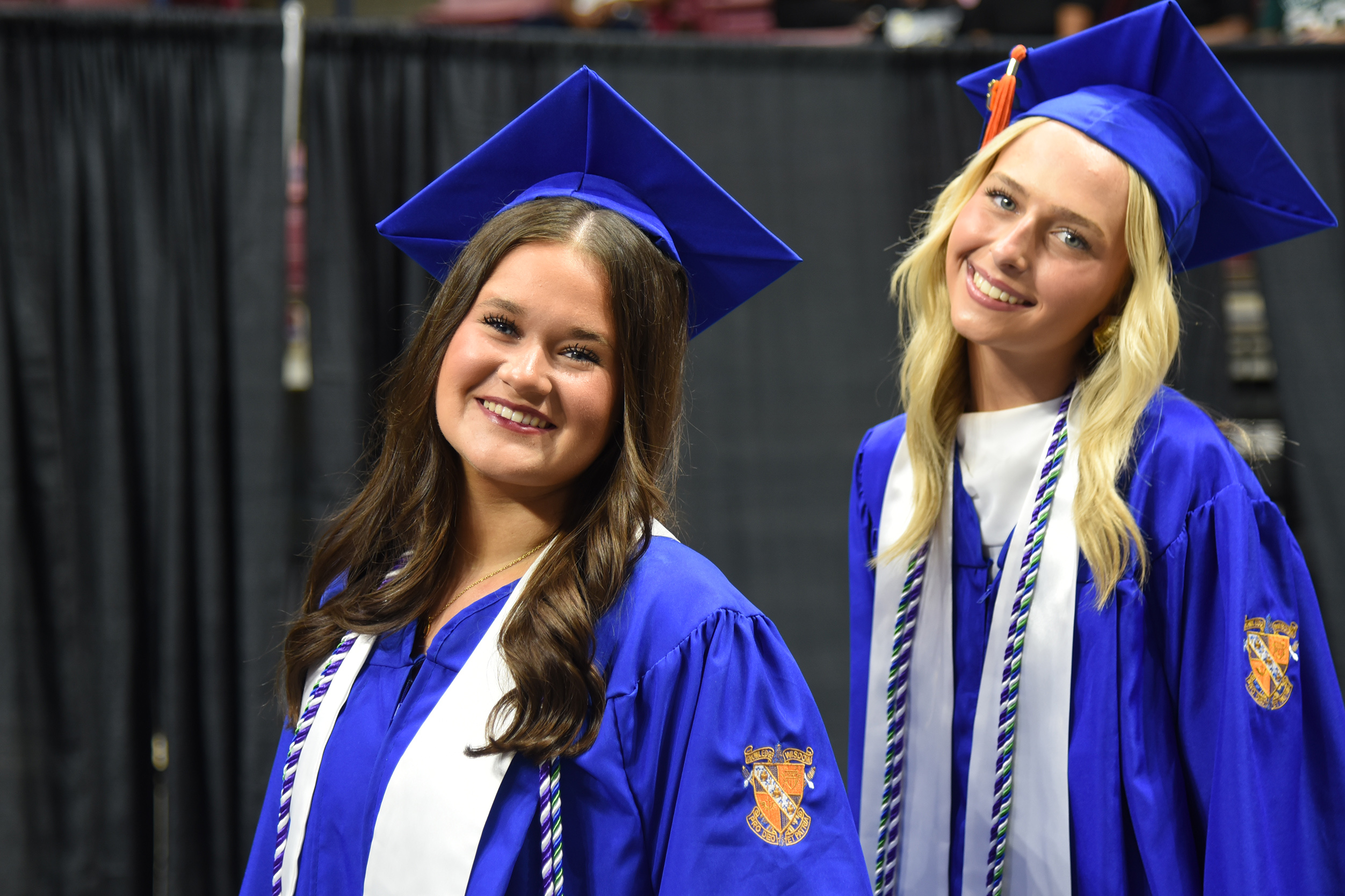 Two young women in blue and white graduation robes and caps smile at the camera in a venue.
