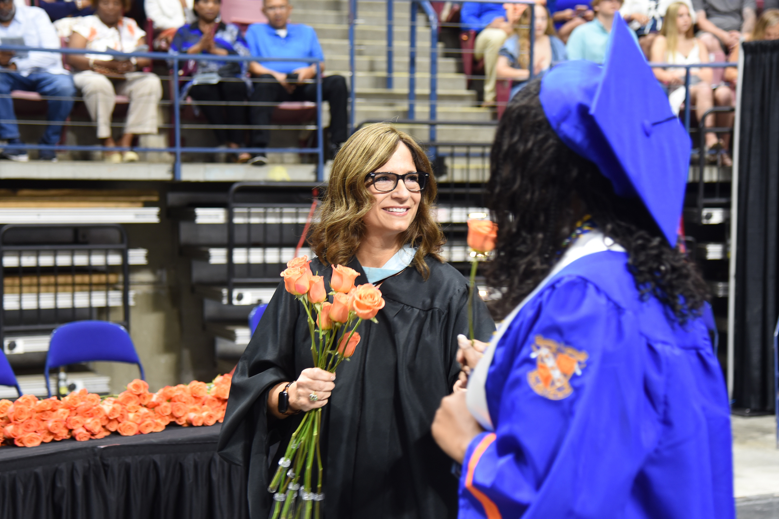 A woman in a black gown and glasses hands flowers to a graduate in a blue gown. Behind them, a crowd watches from bleachers.