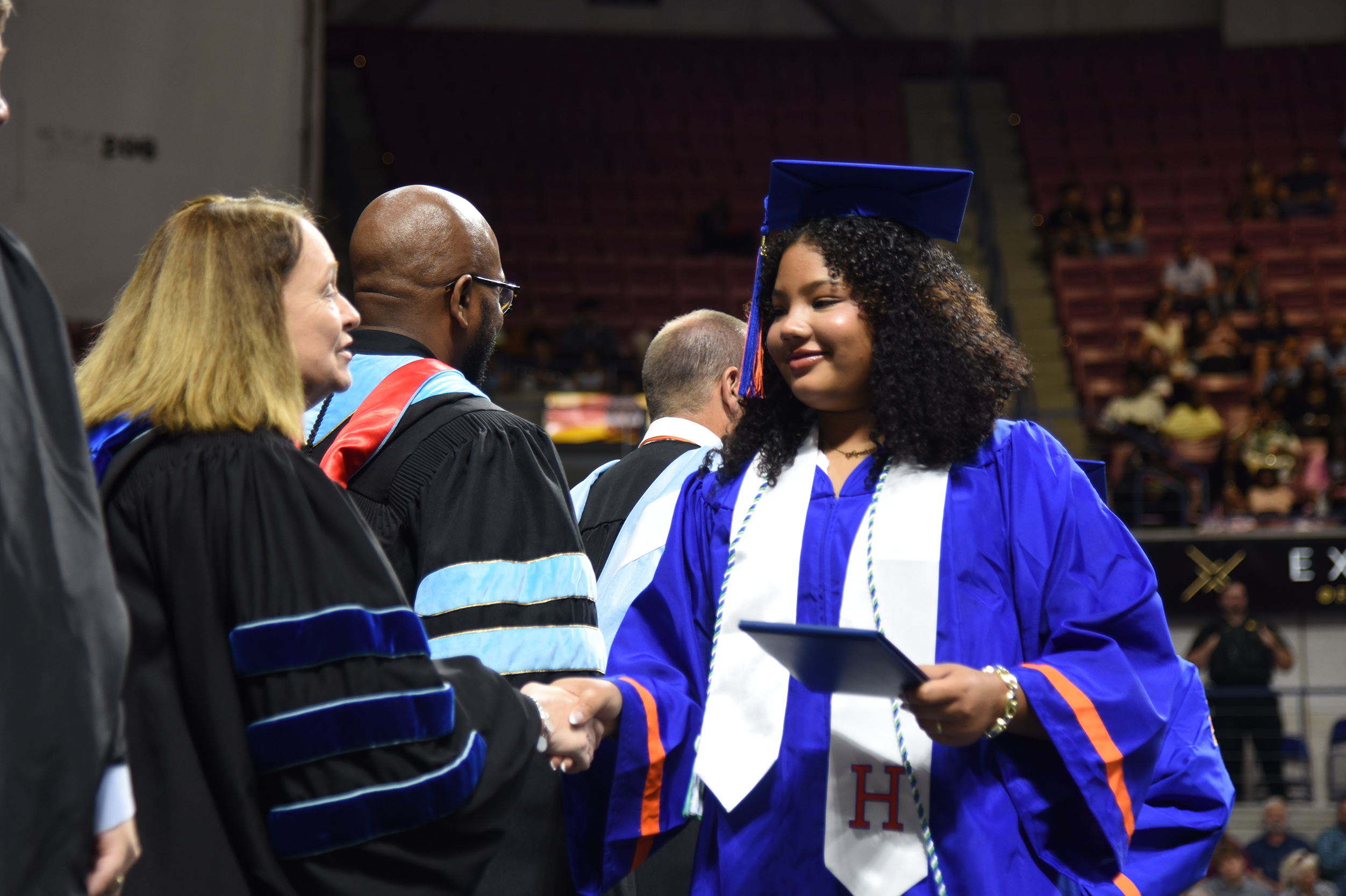 A woman in a blue and white graduation gown shakes hands with a faculty member. Both wear caps.