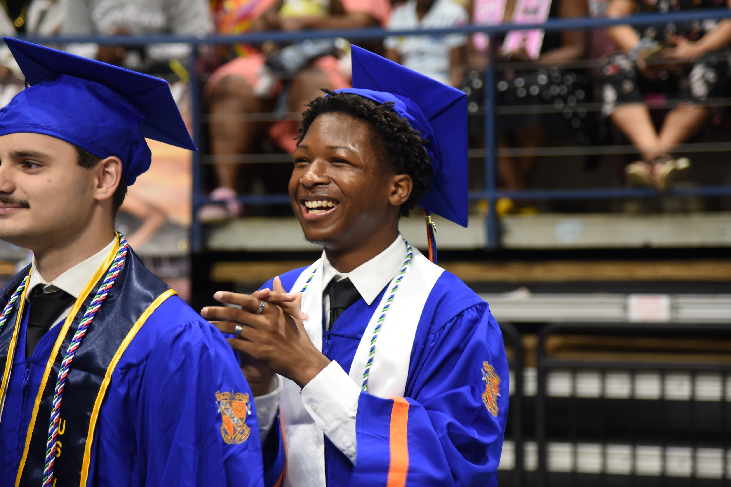 Two men in blue graduation robes and caps stand in front of a blurred audience at a graduation.