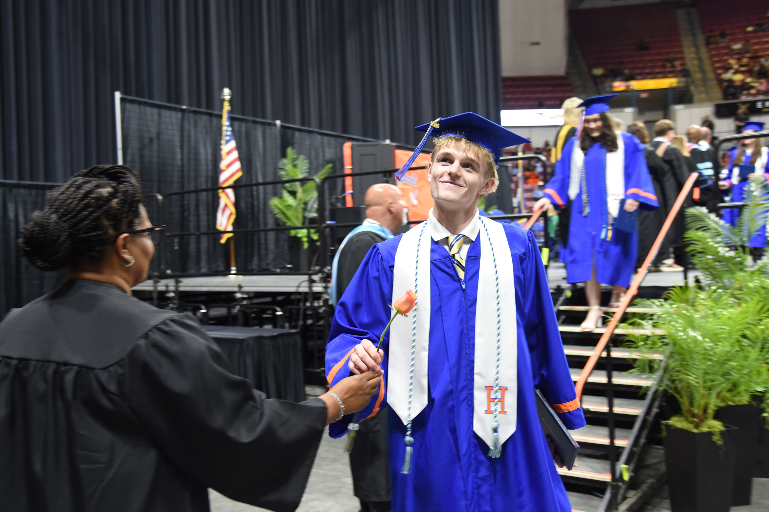 A woman in a black gown hands a rose to a male graduate in a blue gown and cap.