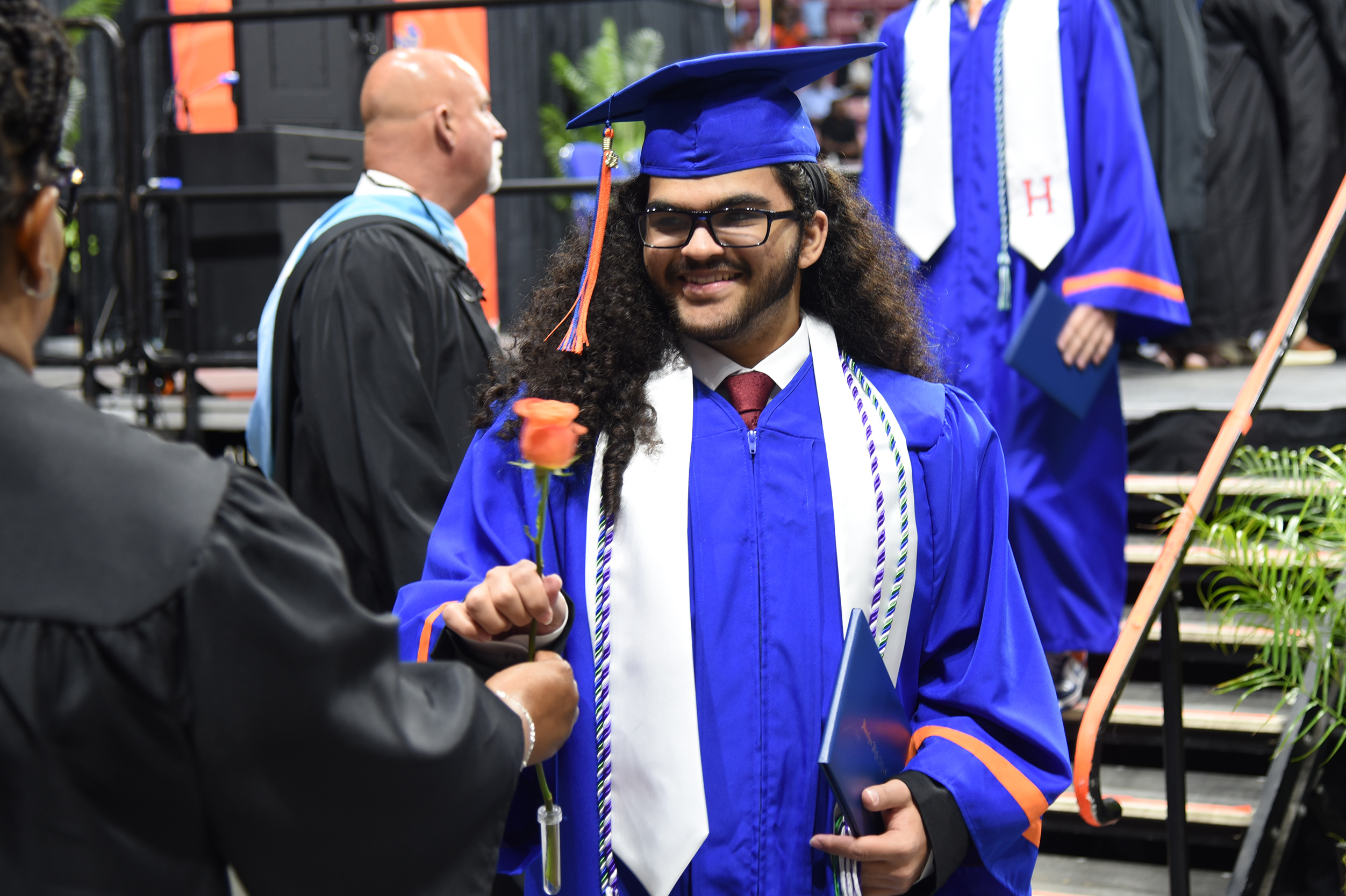 A graduate in blue gown holding a rose receives congratulations at a graduation ceremony.